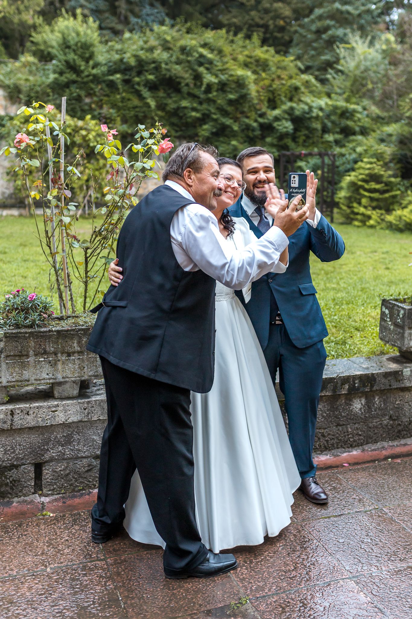 Three people in formal wedding attire share a joyful moment while taking a selfie in a garden setting.