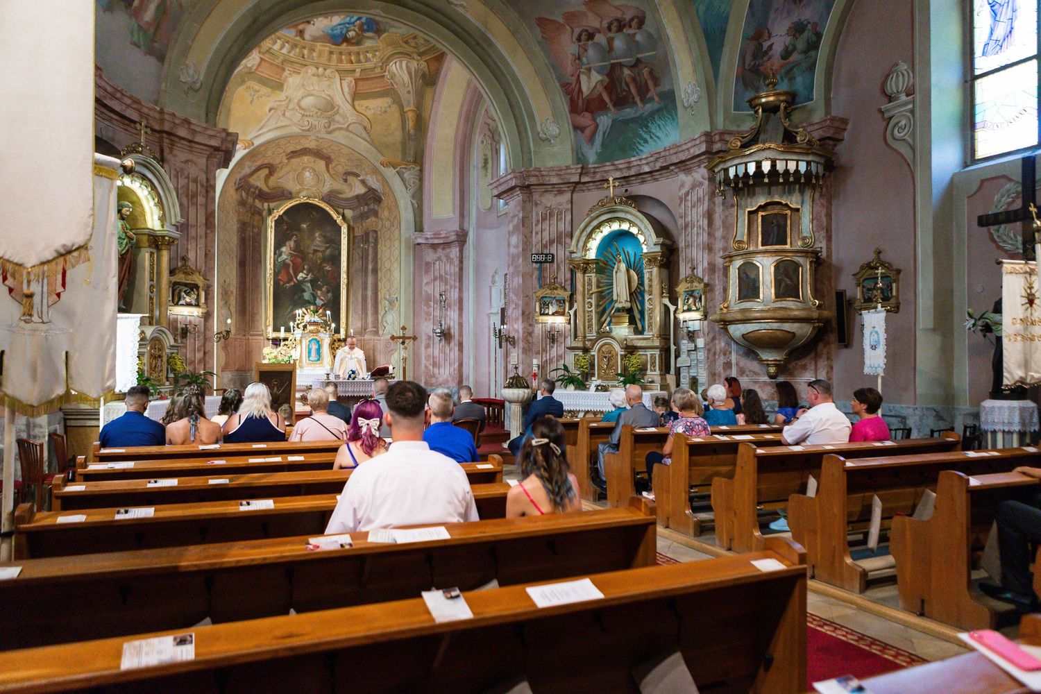 Worshippers sit in wooden pews inside an ornate Catholic church with painted walls and arched ceilings.