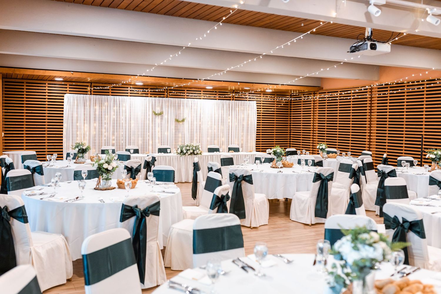 A large banquet hall decorated with white tablecloths and teal accents features elegant table settings and string lights.