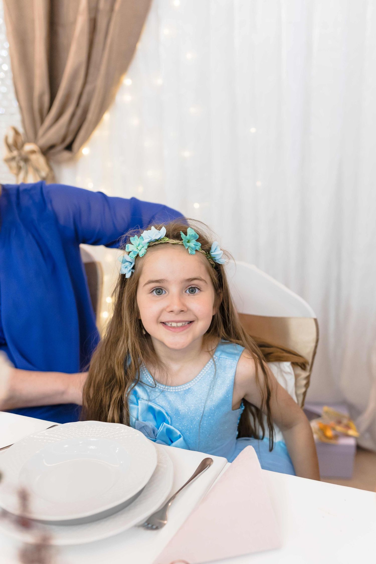 A child in a blue dress and floral crown sits at a table with white plates.