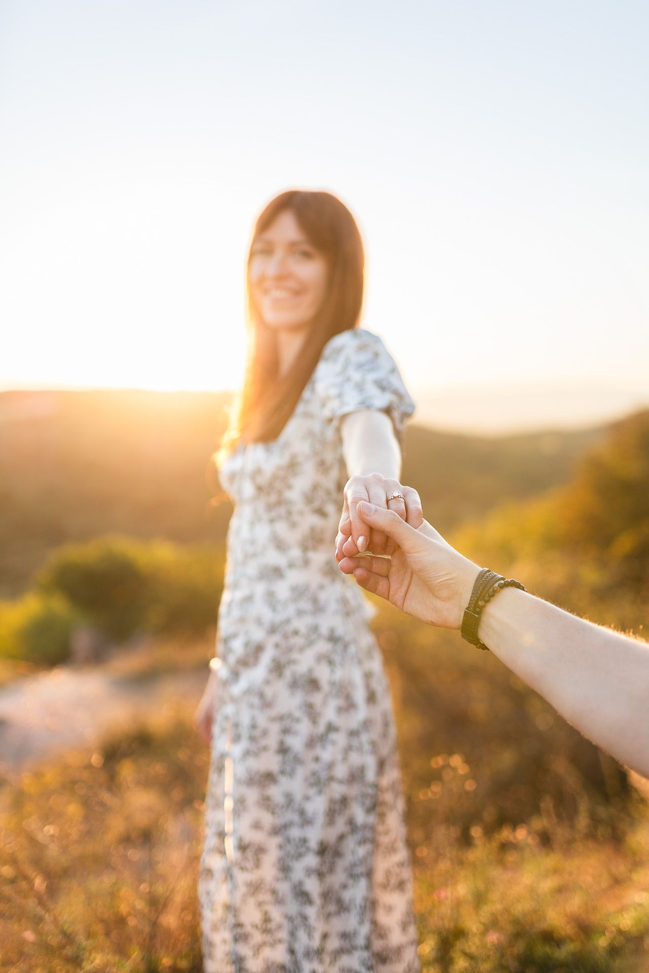 Woman in floral dress twirls joyfully in golden sunlight while showing engagement ring.