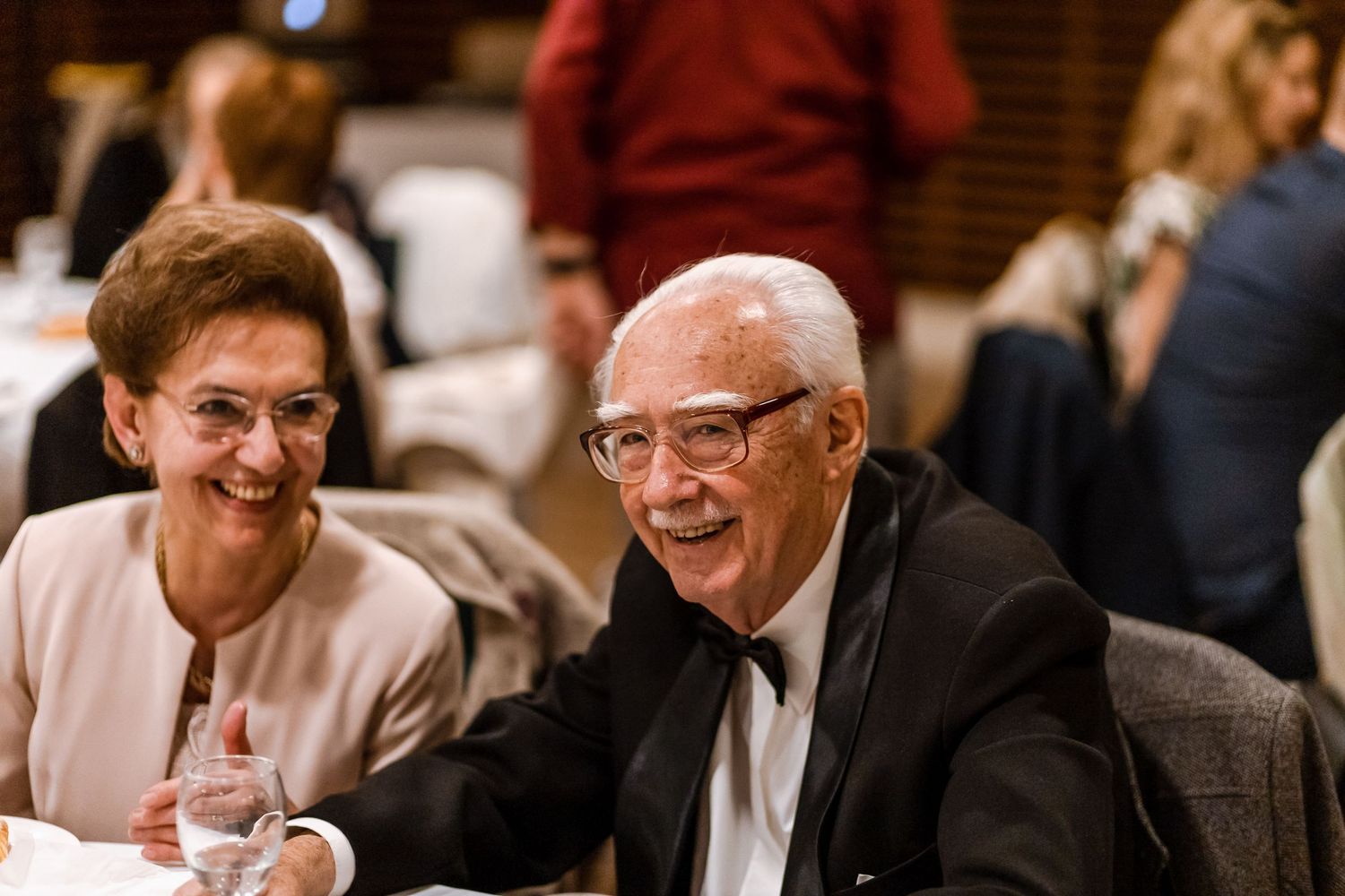A group of people in business attire sit together at a formal event, smiling and engaged in conversation.