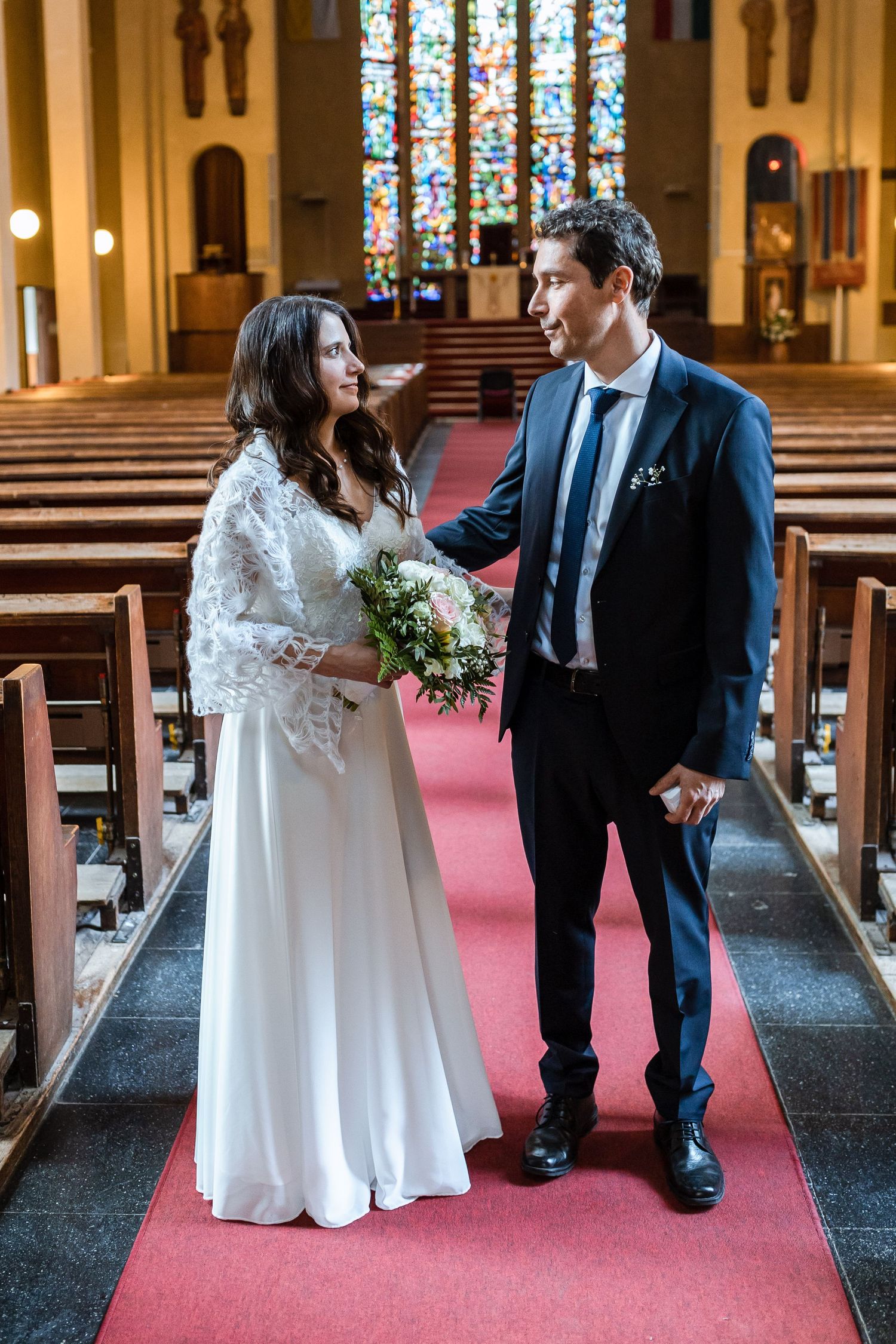 A wedding couple shares a romantic moment in the church aisle with red carpeting and stained glass windows.