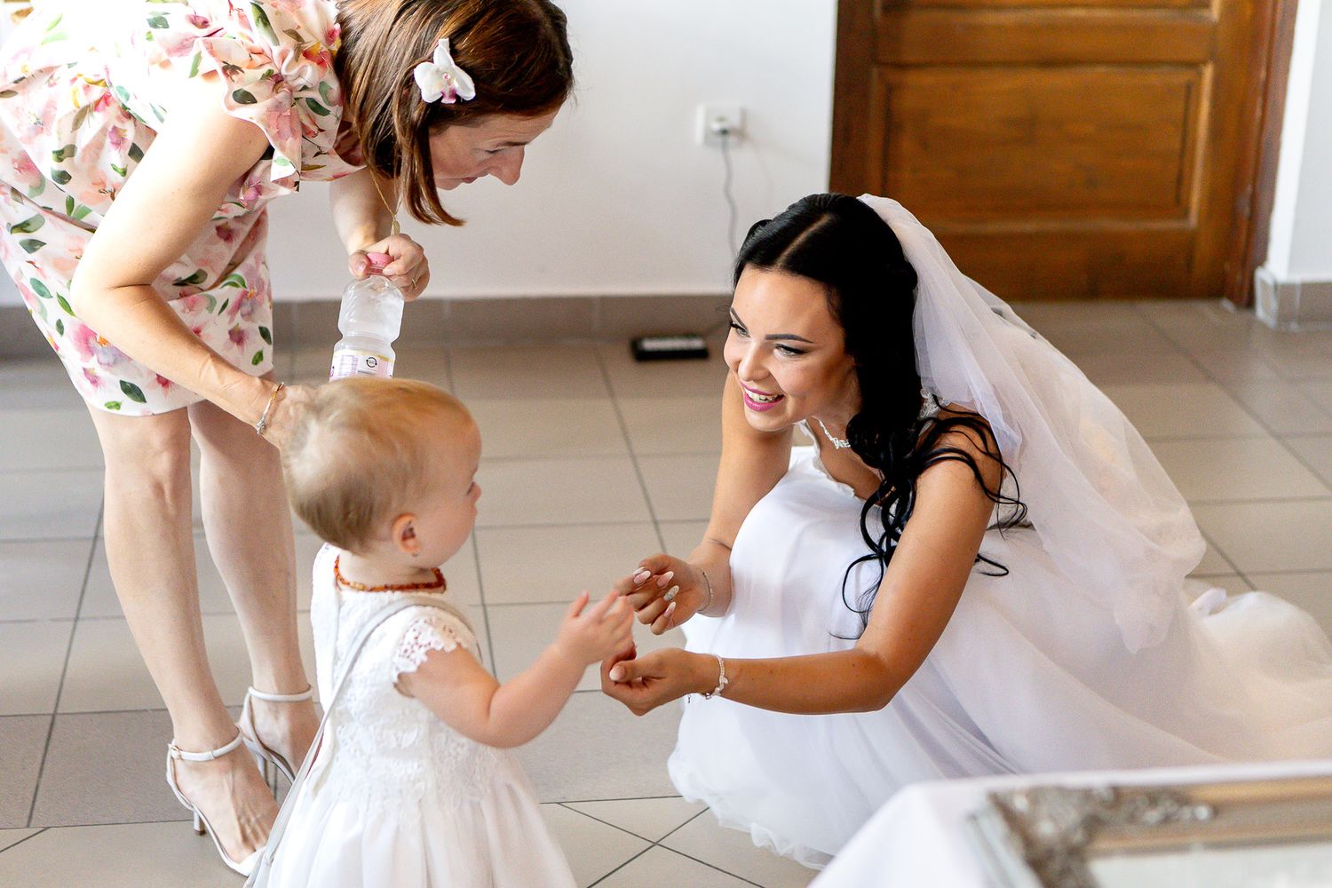 A person in a white dress helps a small child in a white dress with their shoes on a tiled floor.