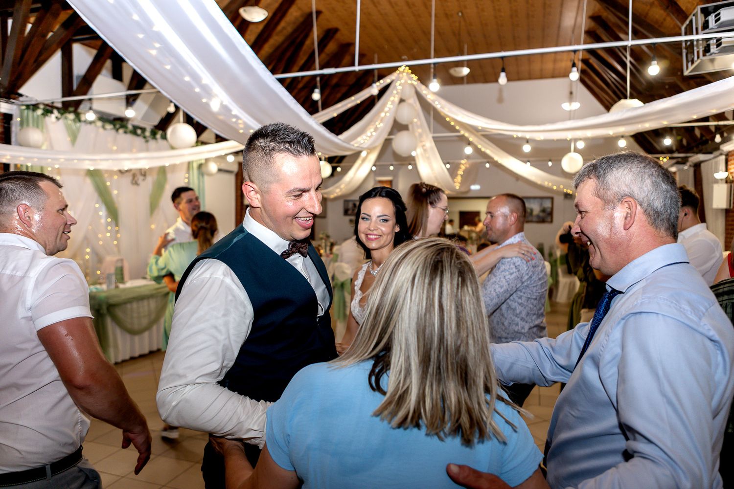 Wedding guests dance and celebrate together under string lights at an indoor reception venue.