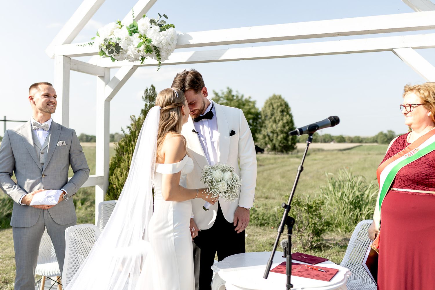 Wedding ceremony guests gather outdoors under a white canopy structure on a sunny day.