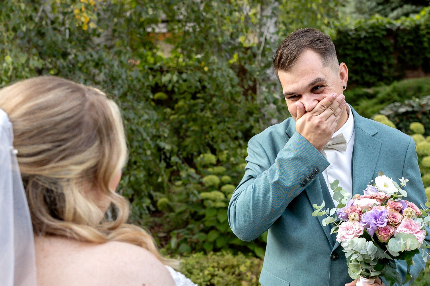 Wedding party celebrates outdoors with joyful reactions amid green garden scenery and colorful floral bouquets.