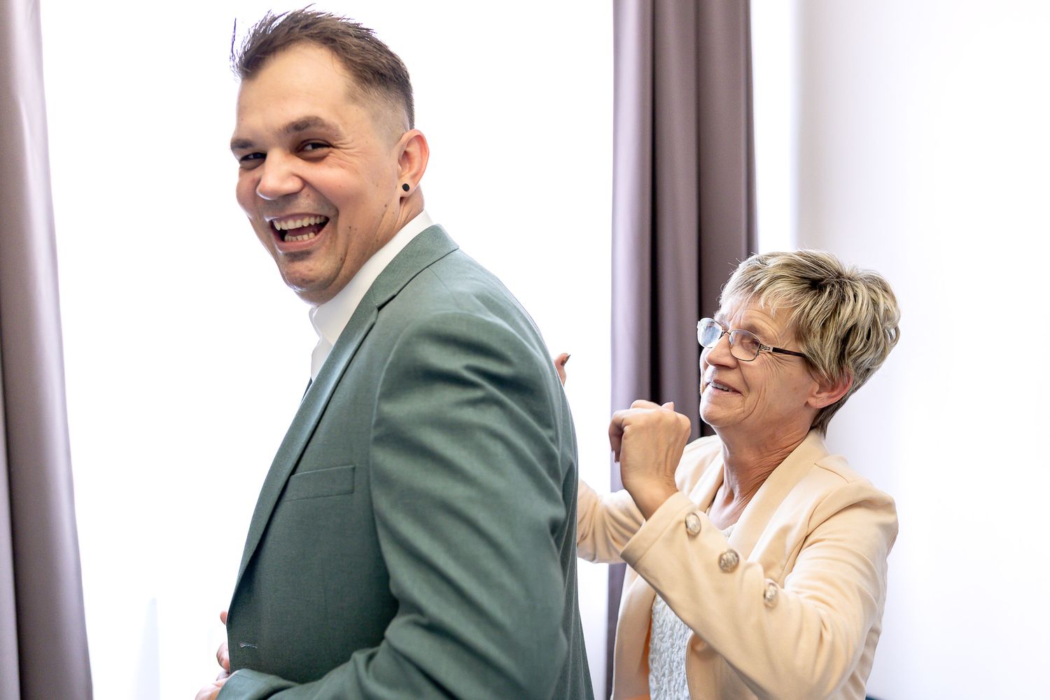 A person in a green blazer and someone in a cream outfit share joyful laughter during an indoor event.