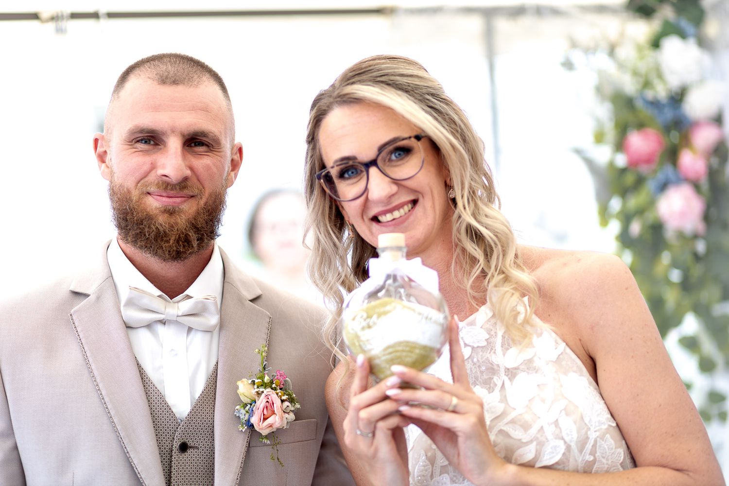 Wedding party in formal attire poses together outdoors with floral decorations and greenery in the background.