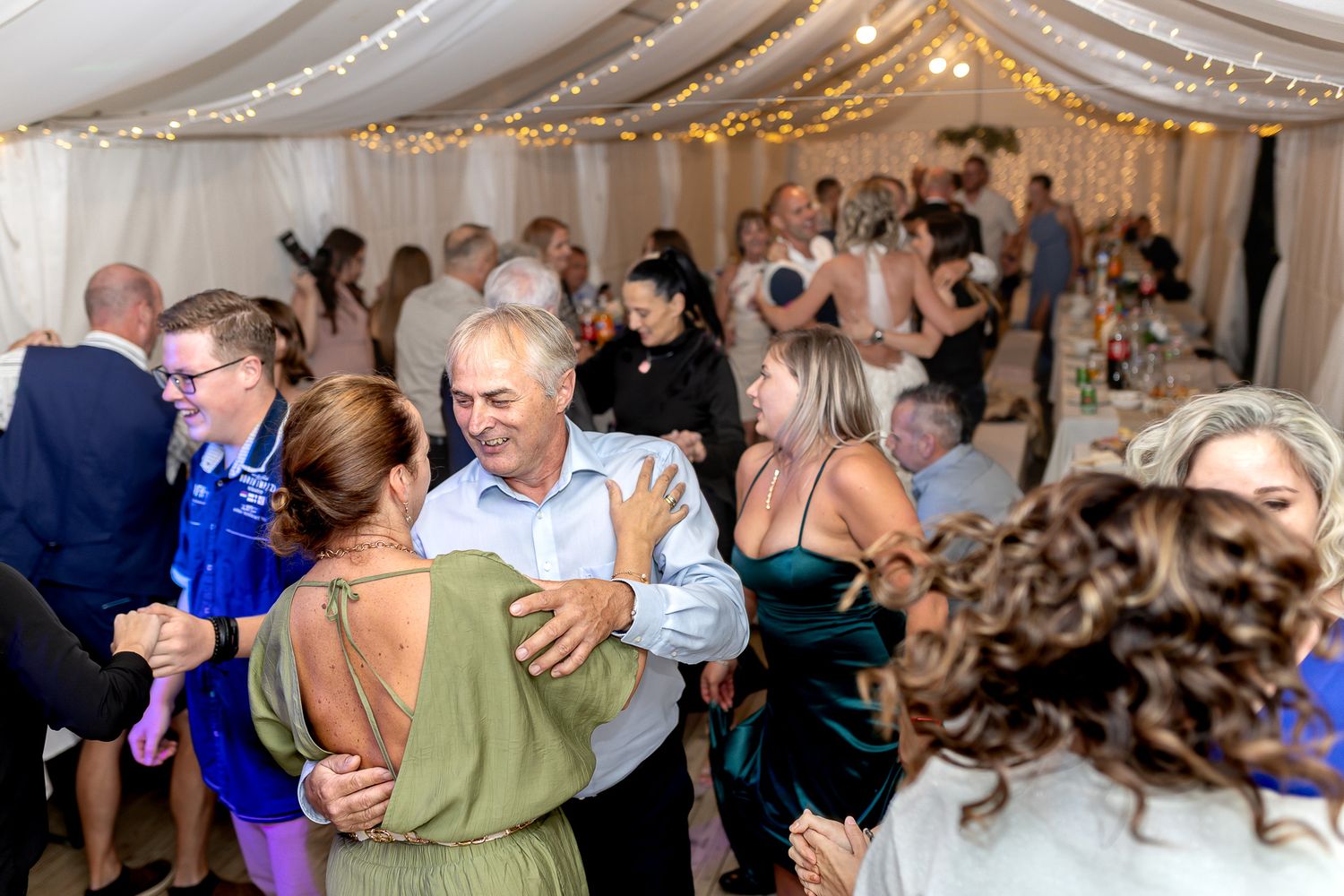 Guests dancing together at an elegant wedding reception with white draped ceiling and warm lighting.