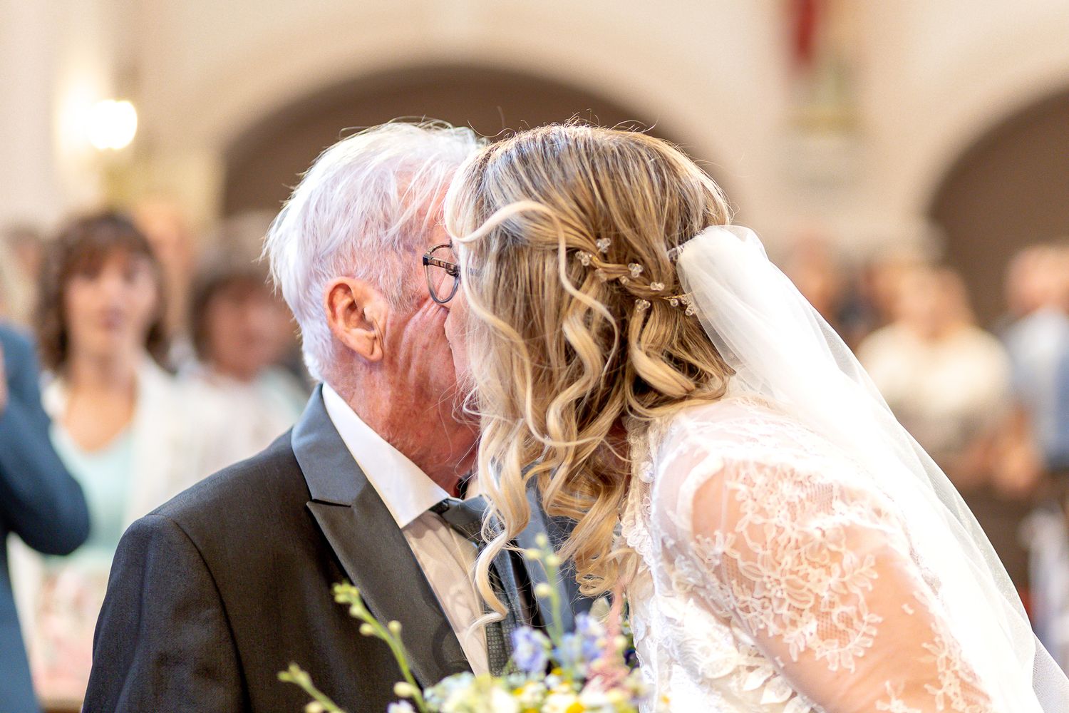 A couple shares a romantic kiss during their wedding ceremony while guests look on in a sunlit venue.