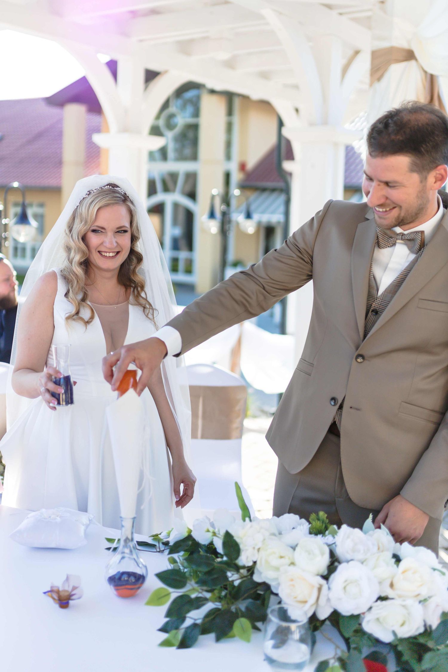 A couple in wedding attire shares a lighthearted moment at an outdoor reception table decorated with white flowers.