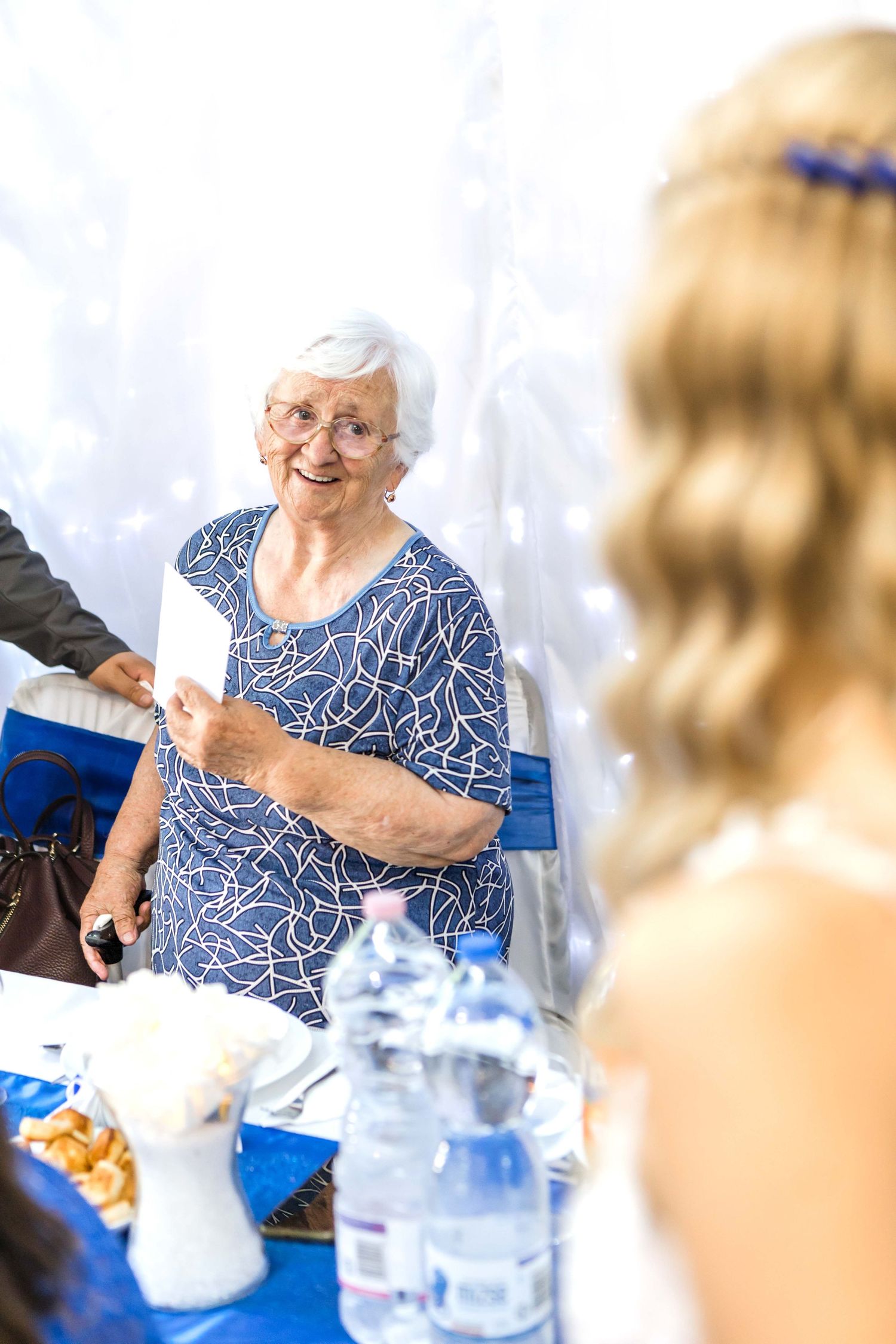 Senior in a blue patterned top sits at a dining table with water glasses and decorative settings.