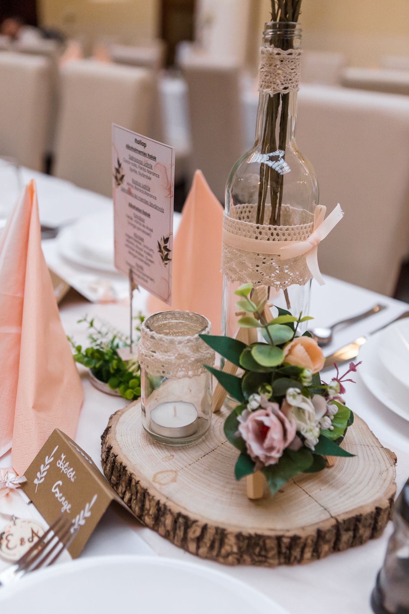 Rustic wedding table centerpiece with wood slice, pink napkins, glass bottle, candle holder and floral decorations.
