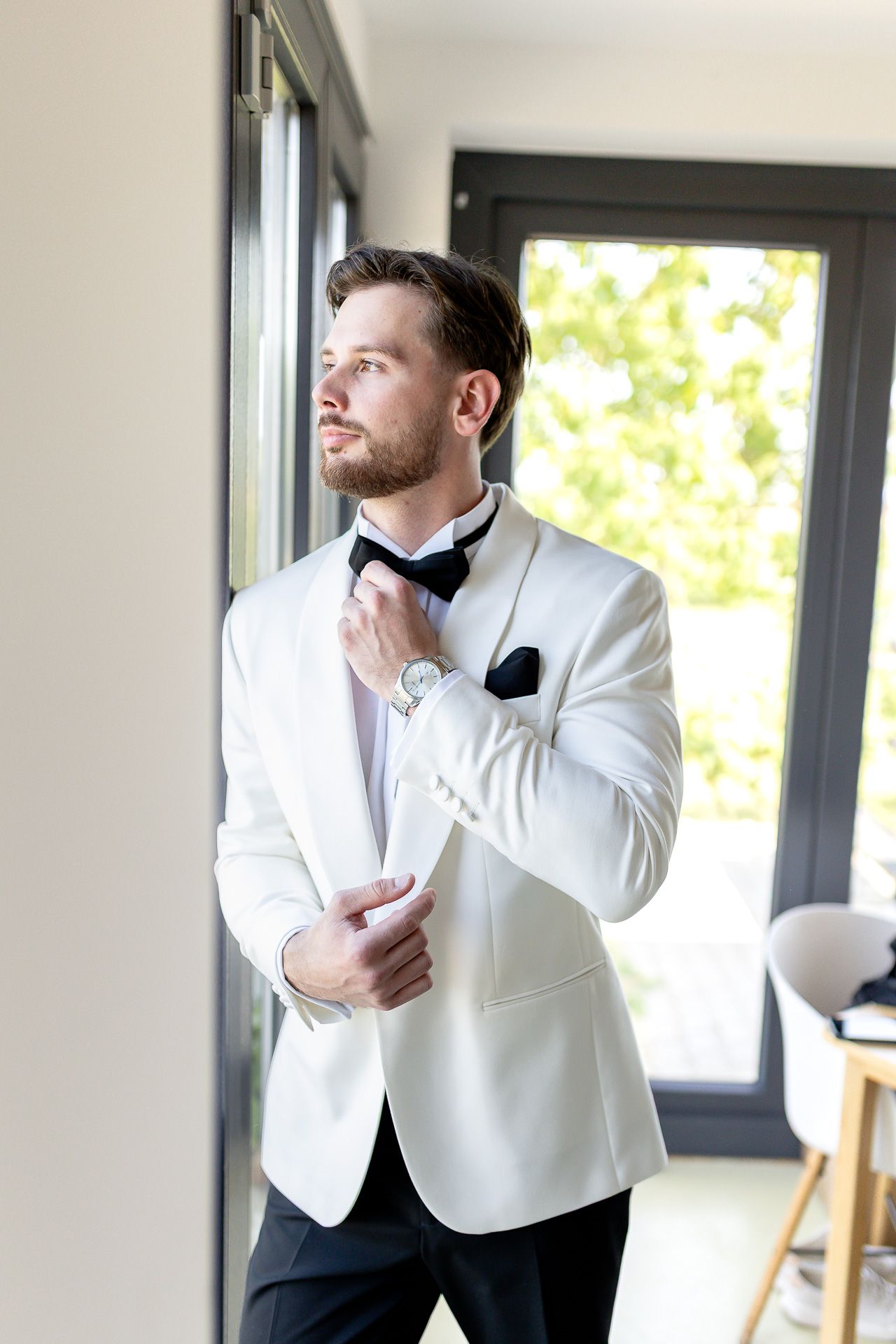 A person in a white tuxedo jacket and black bow tie adjusts their cuff by a window.