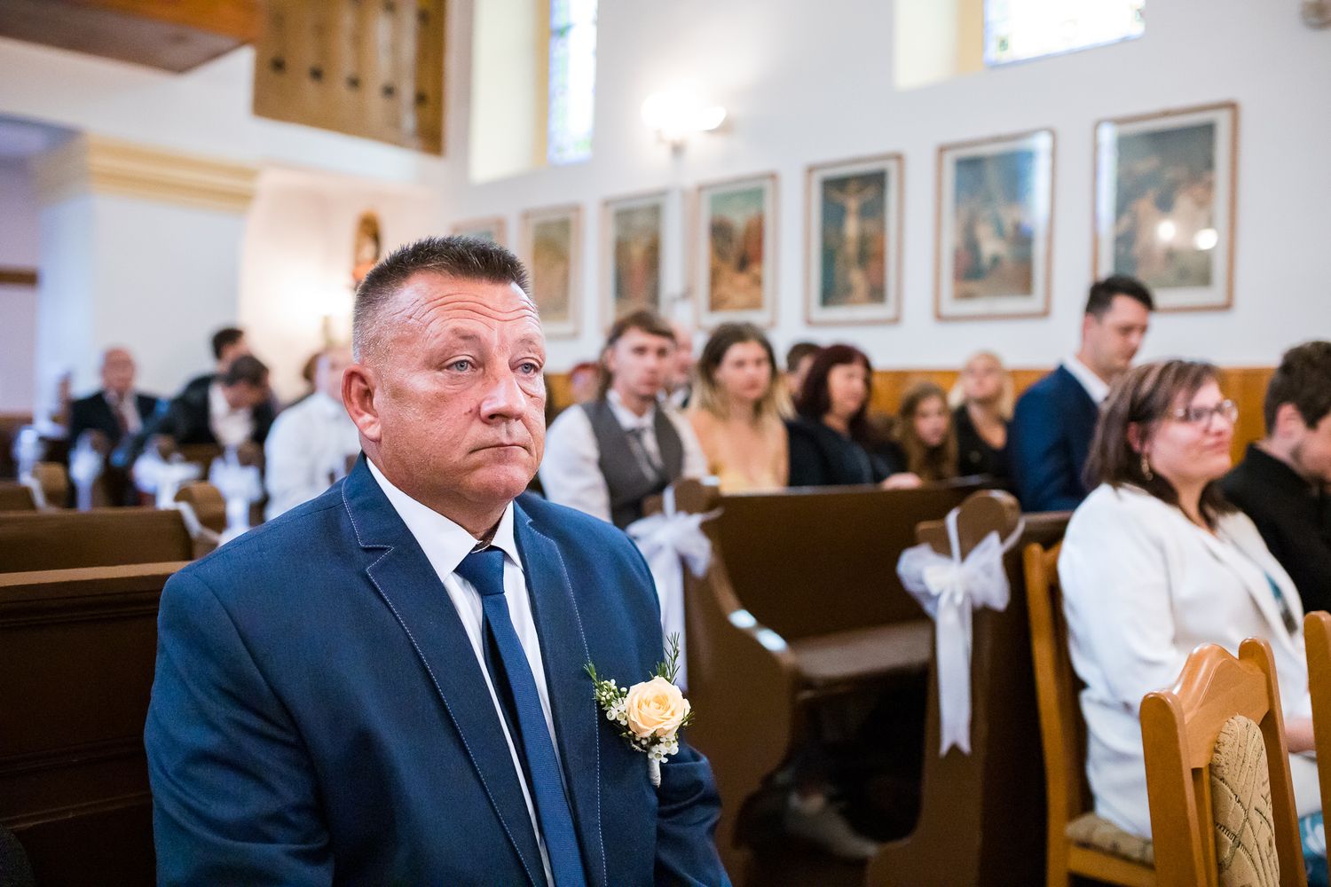 Wedding ceremony guests seated in wooden pews of a traditional church with portrait frames on the wall.