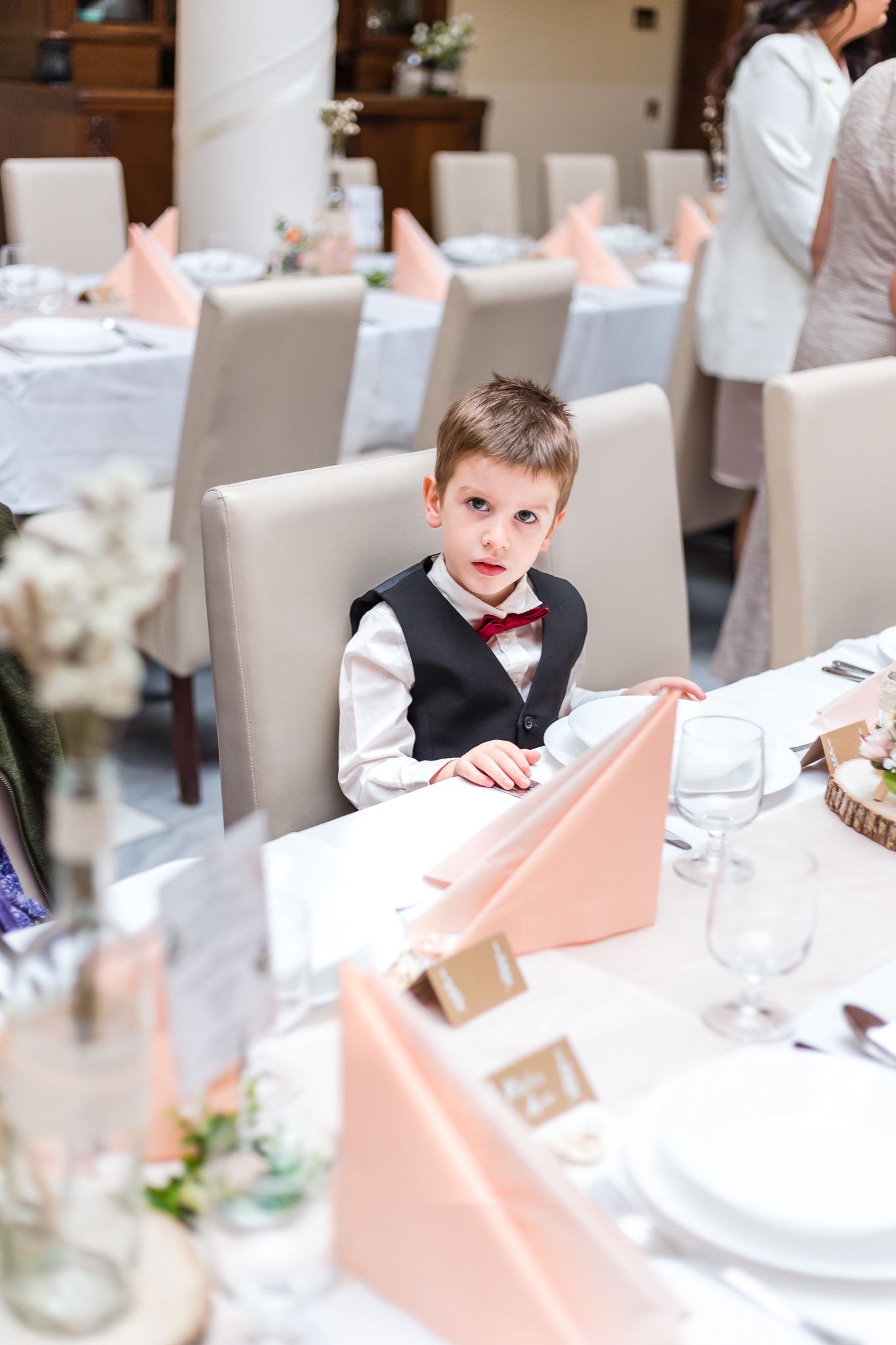 Young boy in white dress shirt and bow tie sits at an elegant table setting with peach colored napkins at formal event.