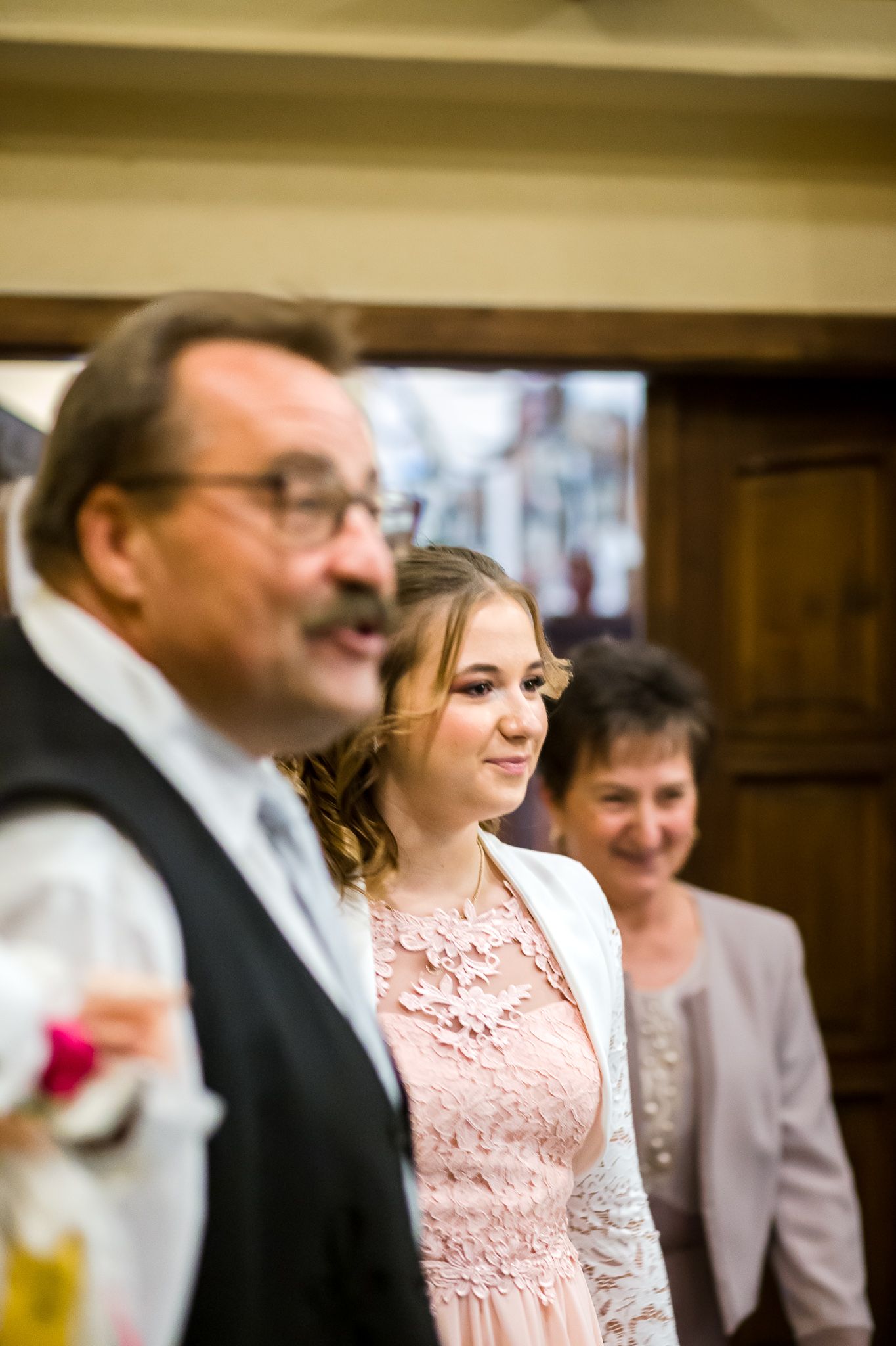 Three people stand together at a formal event, with one wearing a pink lace dress and another in a dark vest.