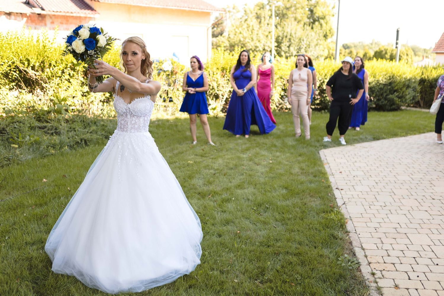 A bride in a white ballgown twirls on a stone pathway surrounded by greenery and guests in blue dresses.
