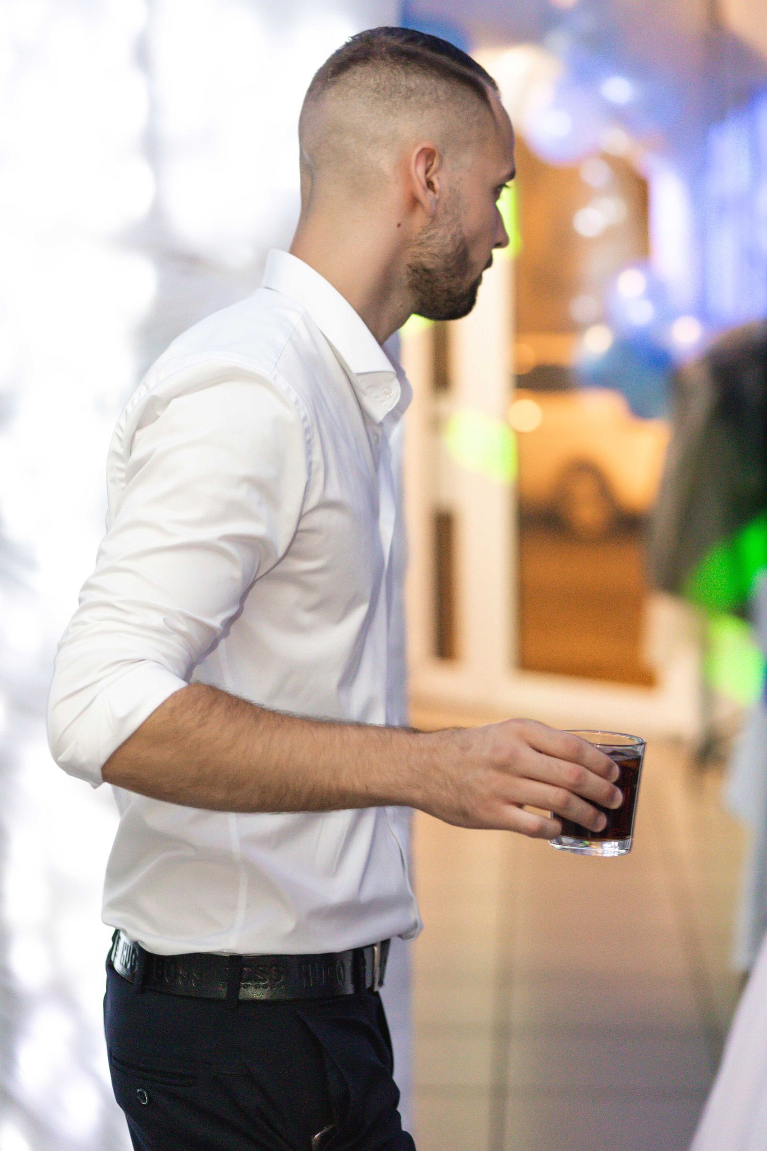 A person in a white dress shirt and dark pants stands in a blurred indoor setting with bokeh lights in background.