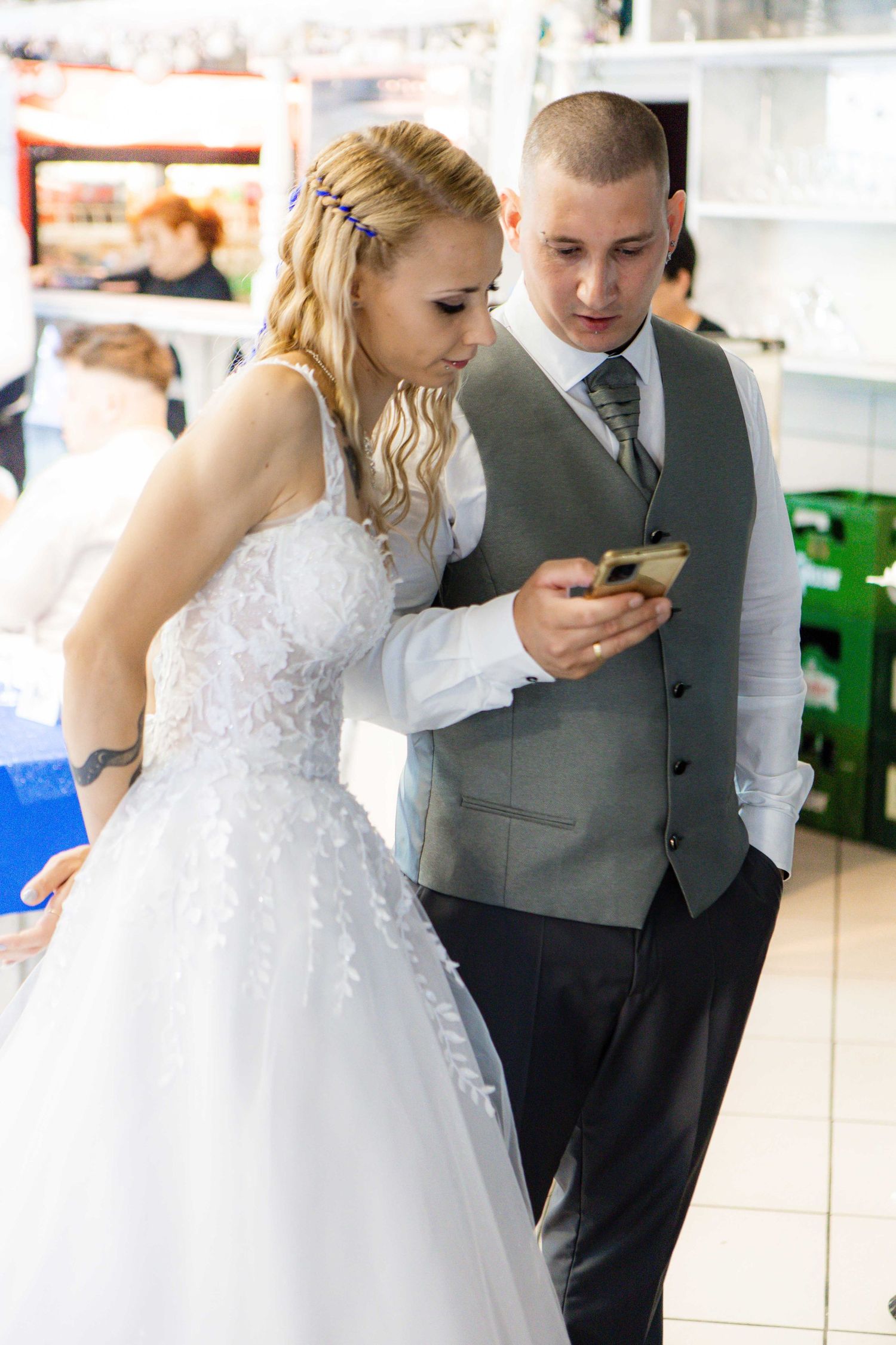 A bride in a white lace gown and groom in a gray vest look at a mobile phone together during their wedding day.