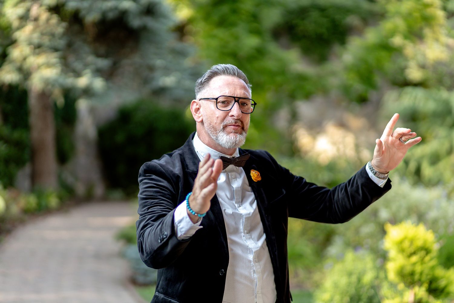 A person in a black suit and glasses gestures expressively with their arms in an outdoor garden setting.