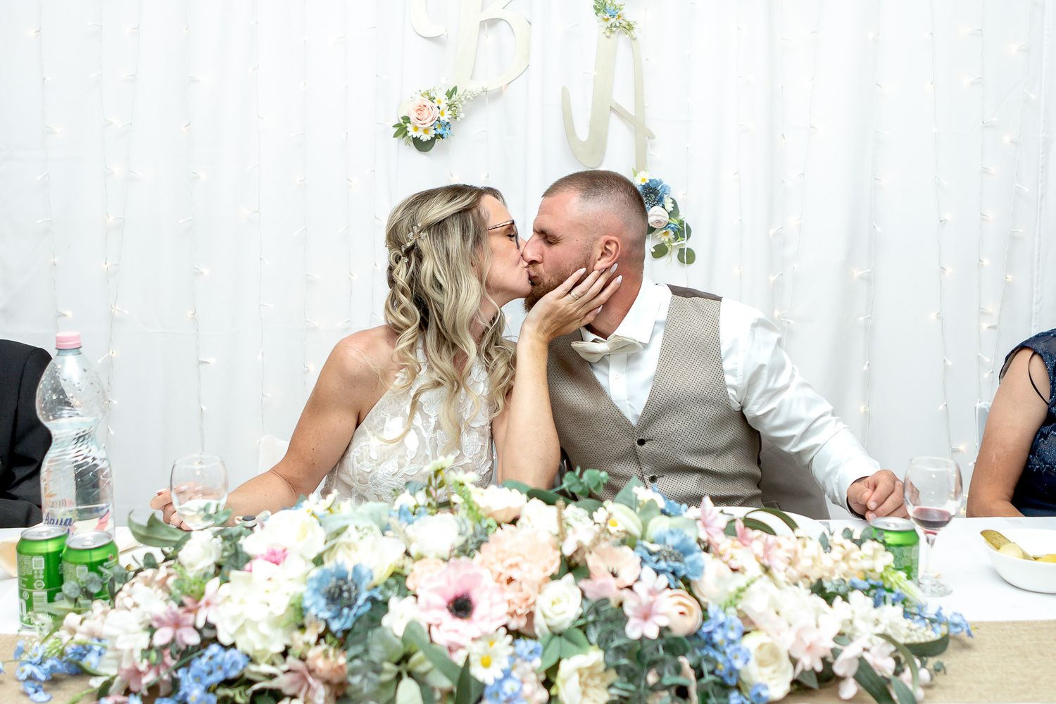 Couple shares a romantic kiss at a wedding reception with floral decorations and twinkle lights in the background.