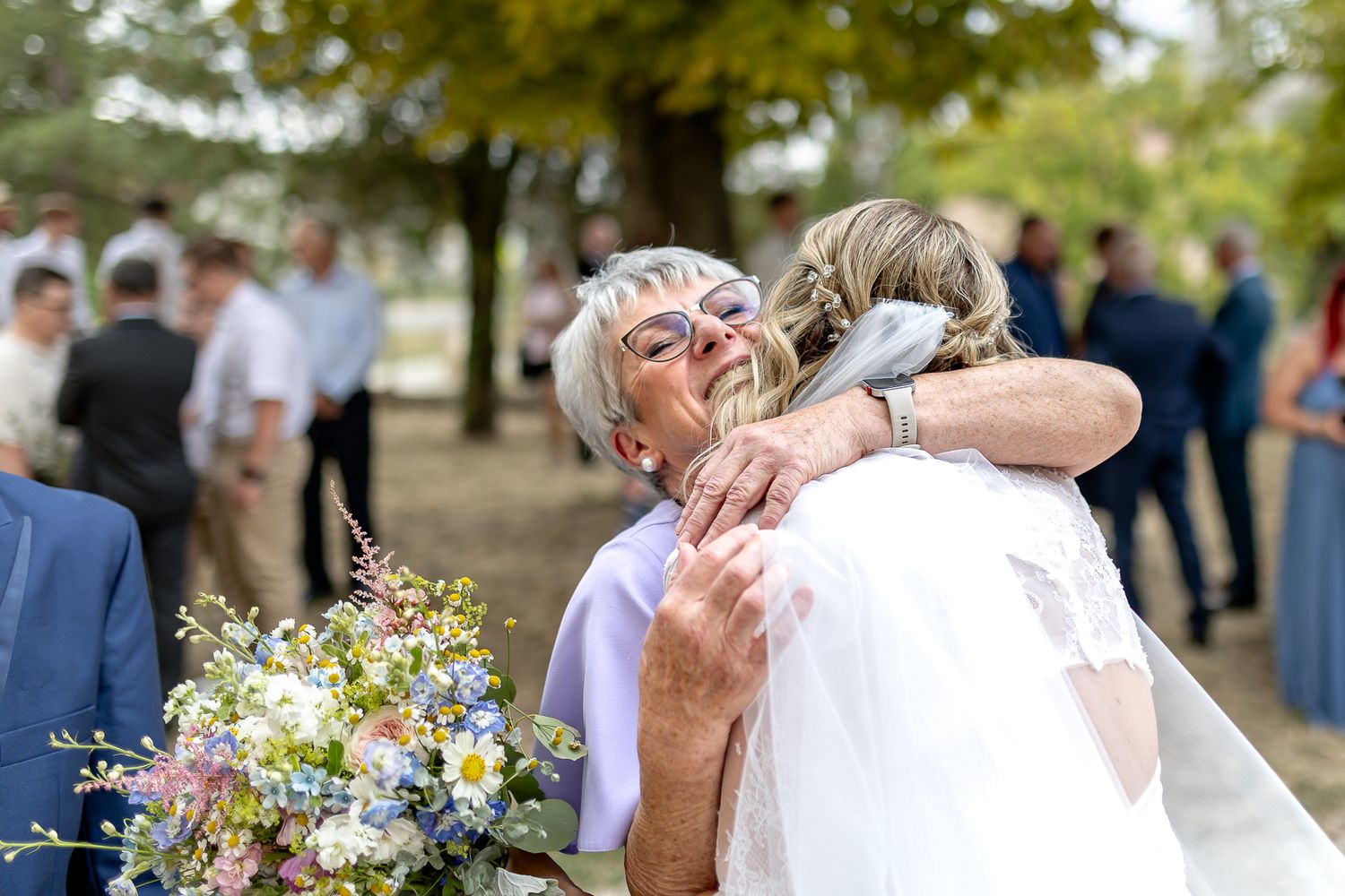 A couple embraces during an outdoor wedding ceremony surrounded by trees and guests in a natural setting.