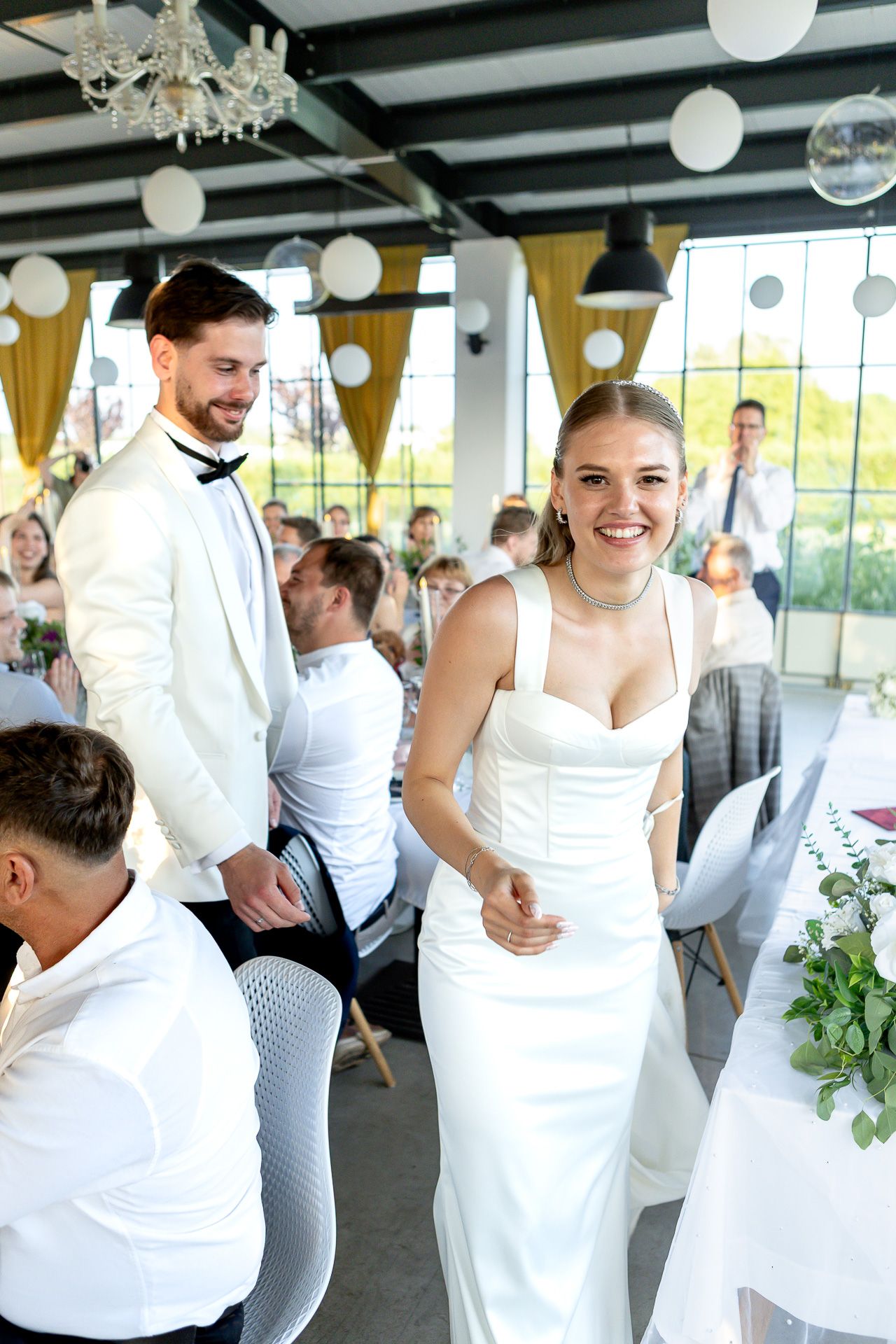 Wedding party members dressed in white gather for a celebration in a modern venue with yellow curtains.
