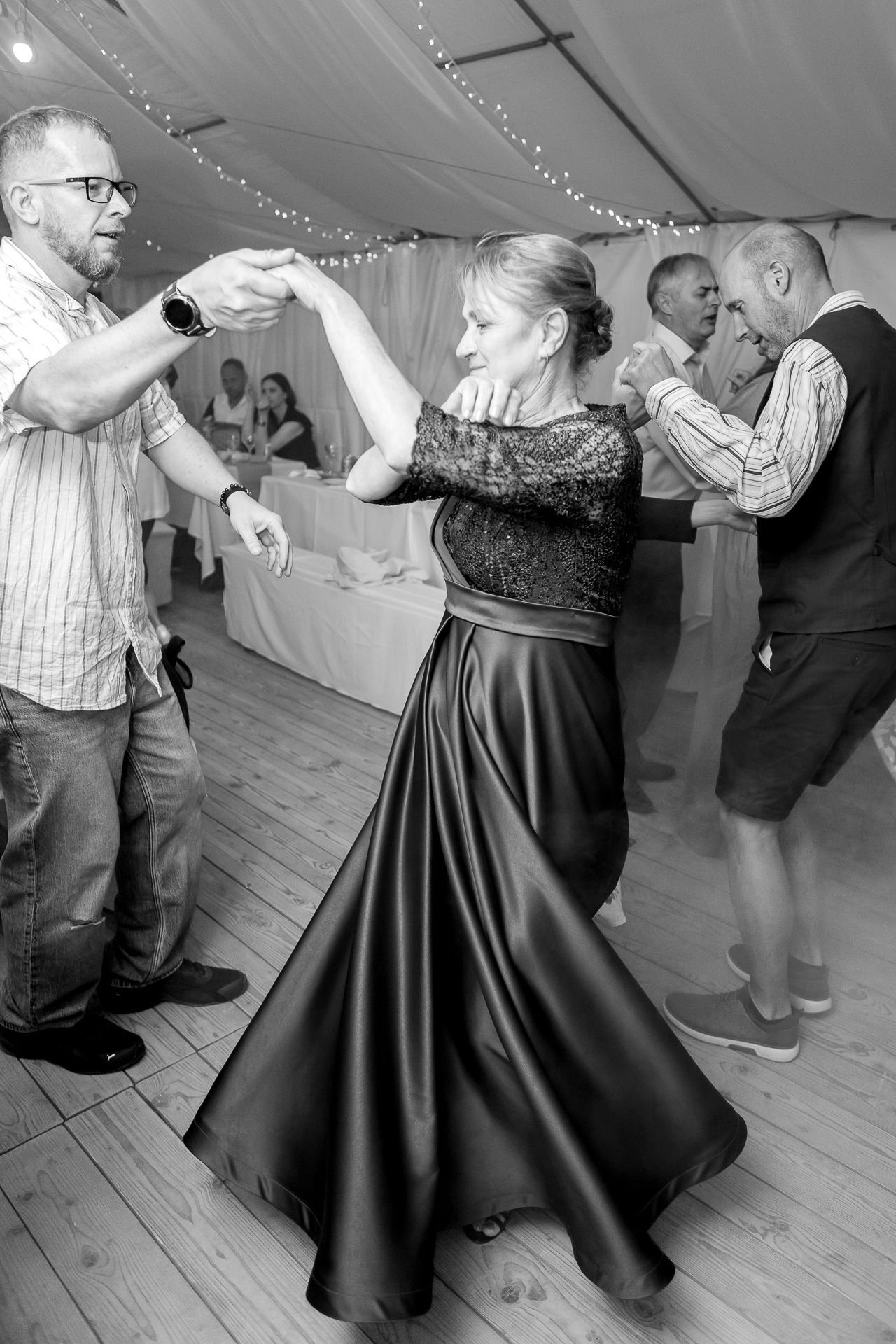Couples dance gracefully across a wooden floor in a tent decorated with twinkling lights during a formal event.