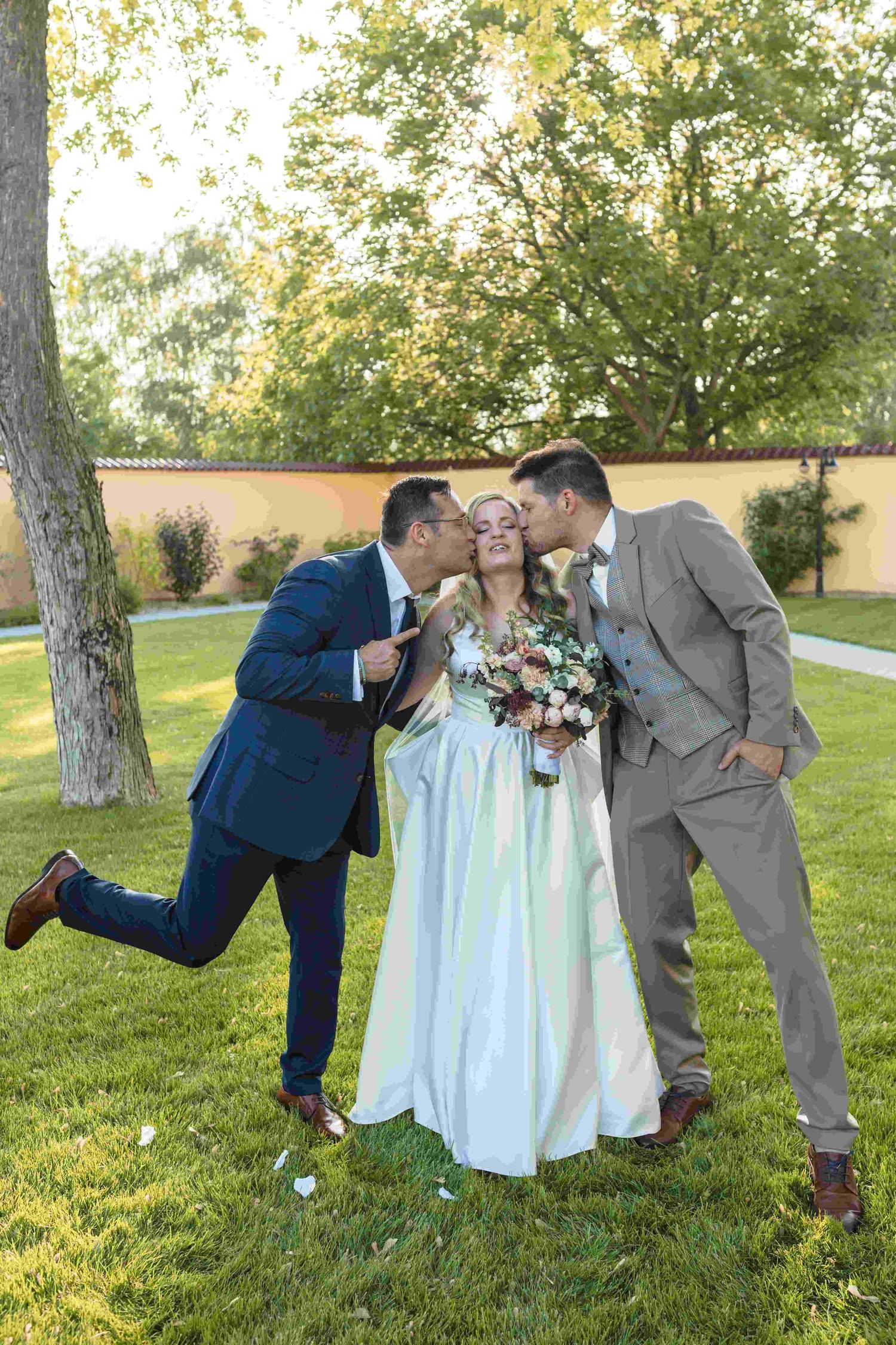 A joyful wedding moment captured outdoors with a bride in white being kissed on both cheeks in a garden setting.