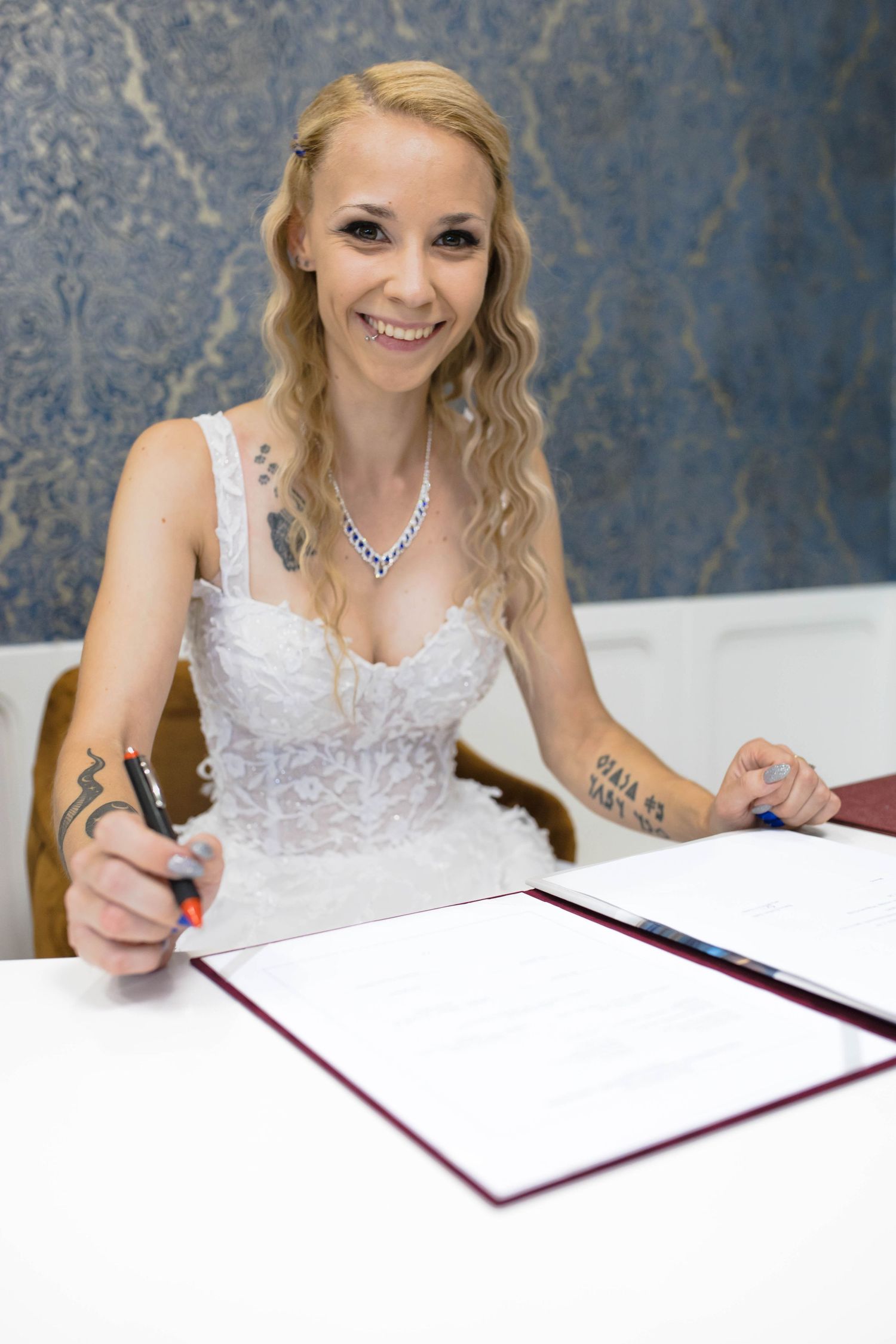 A person in a white lace wedding dress smiles while signing documents at a table.