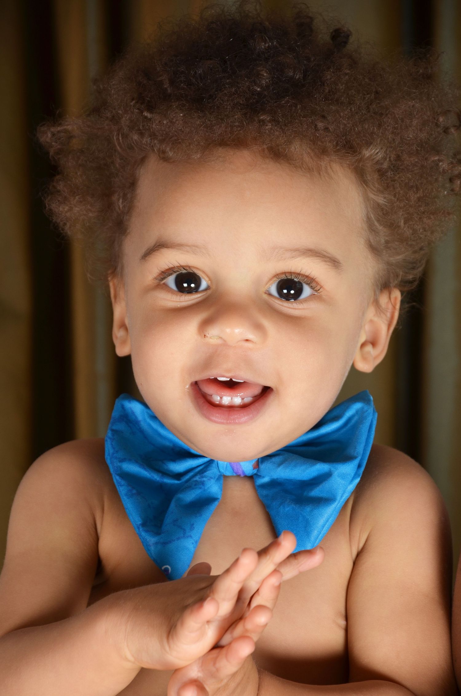 A baby wearing a bright blue bowtie smiles at the camera in a close-up portrait.