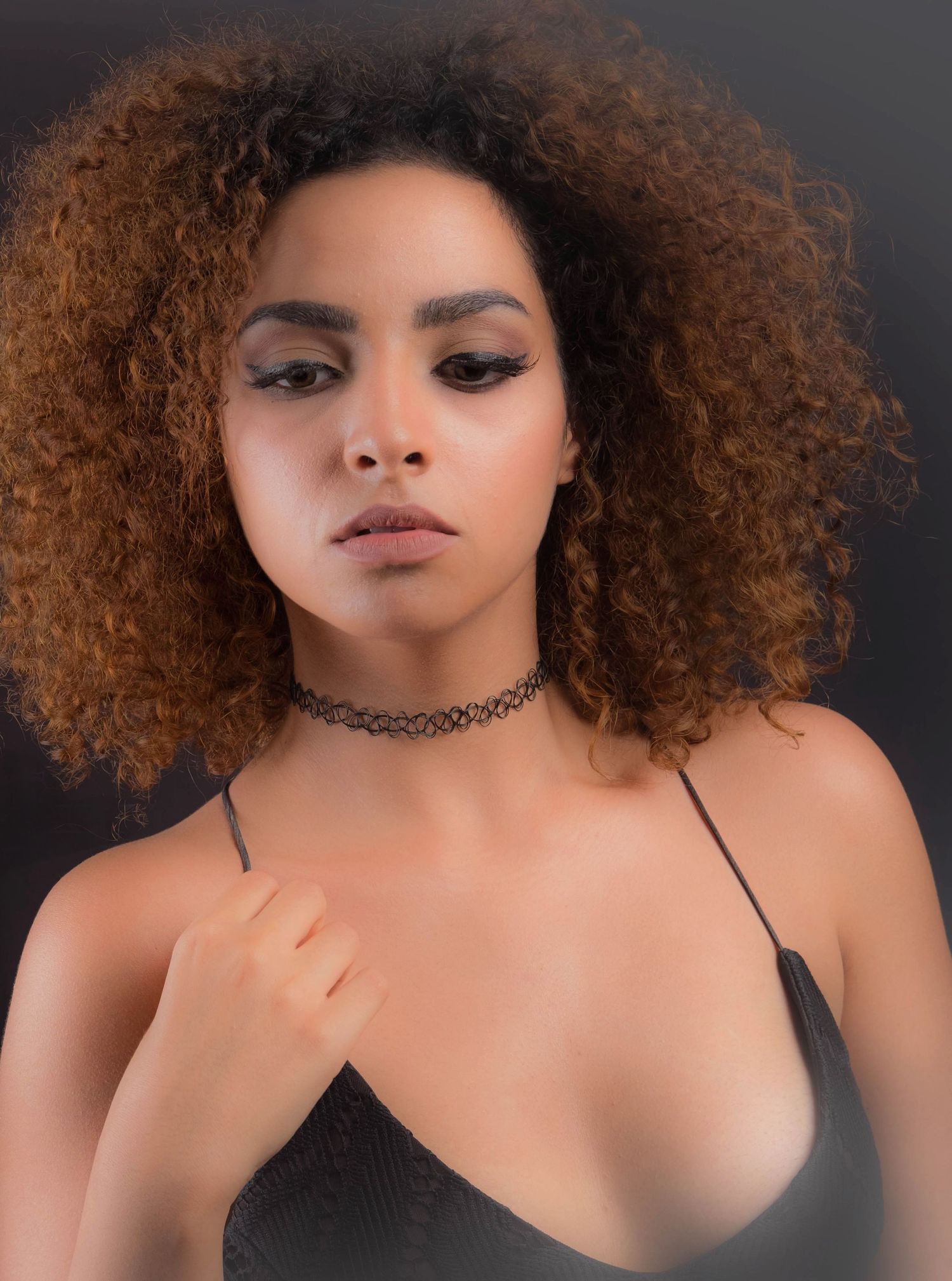 Studio portrait with natural curly hair and choker necklace against dark background.