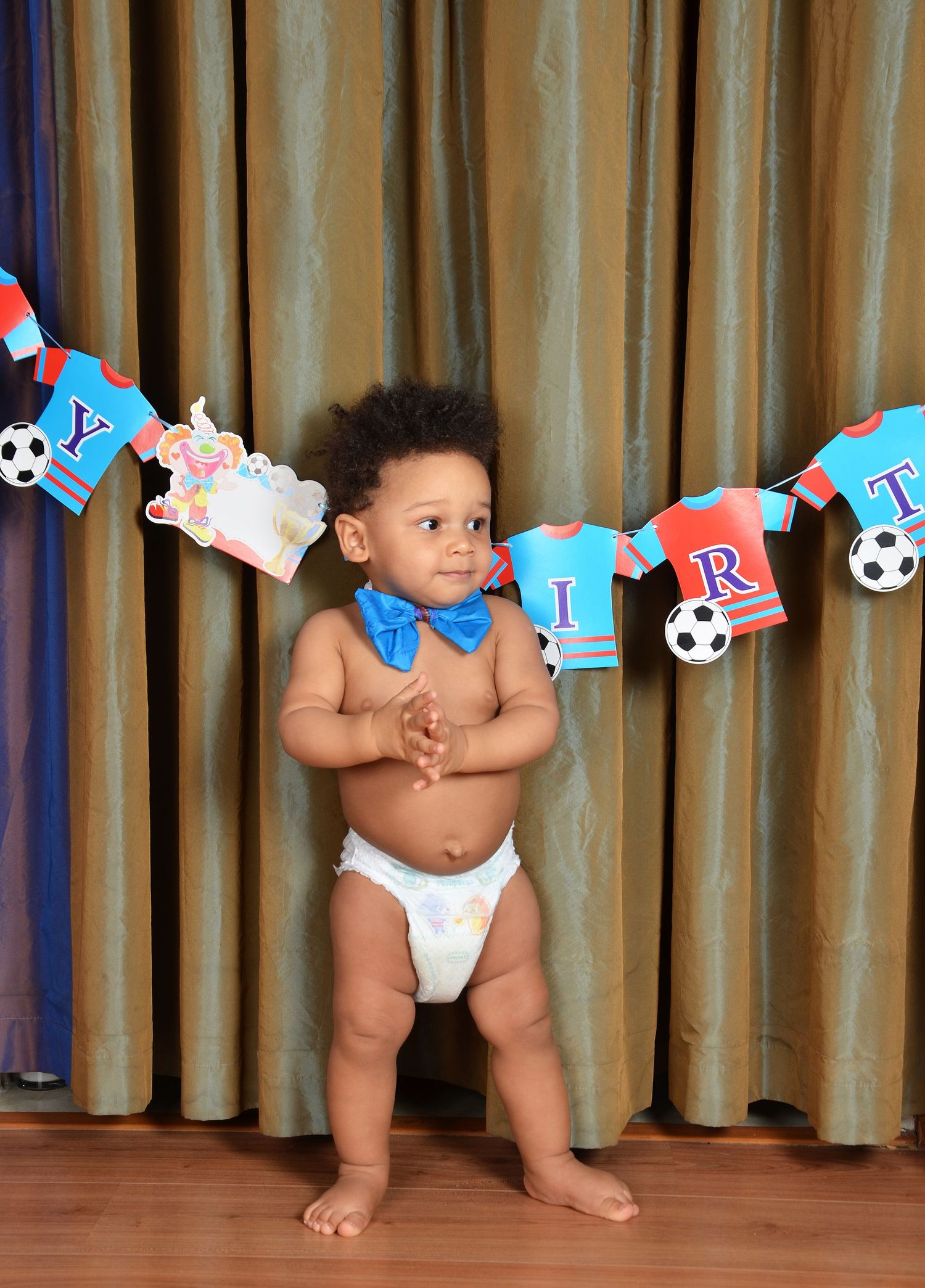 A toddler in a diaper and blue bowtie stands confidently by a birthday banner with soccer decorations.