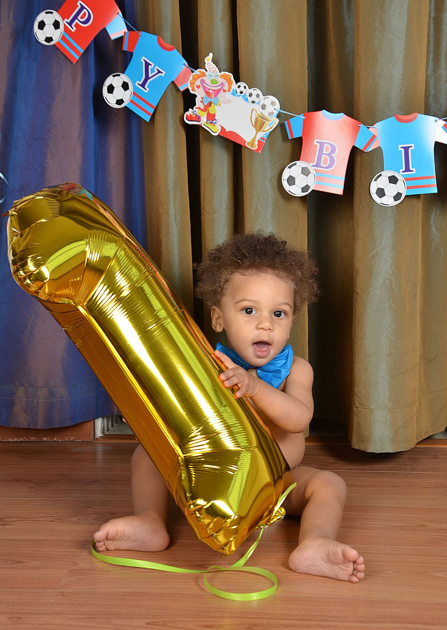 A baby poses with a large gold number 1 balloon beneath a soccer-themed birthday banner.