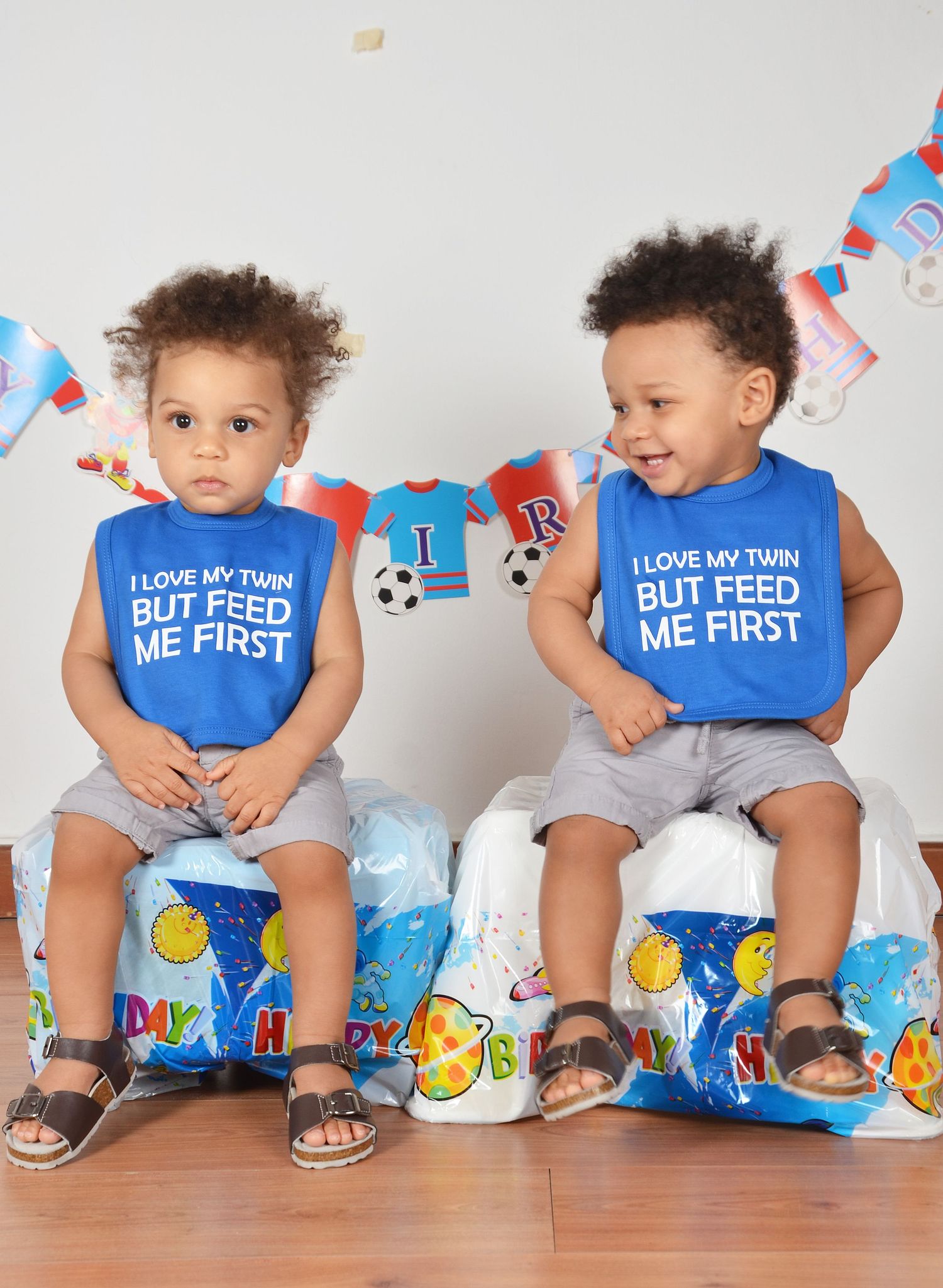 Twin toddlers in matching blue shirts sit on birthday decorations smiling and looking serious.