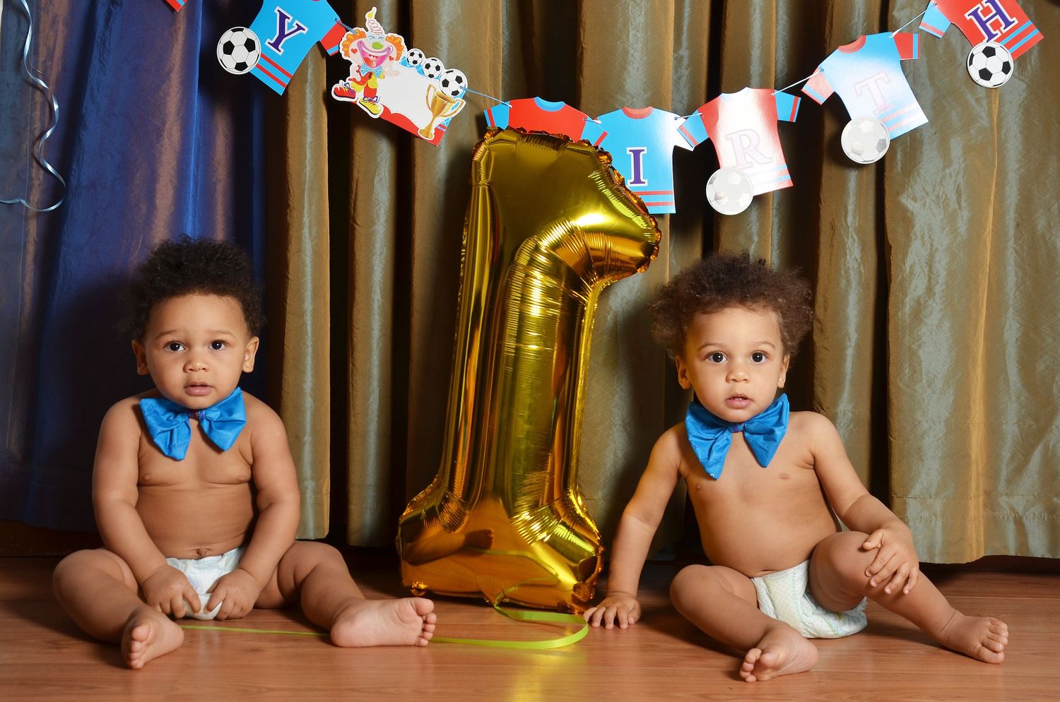 Two babies sit on floor with a gold number 1 balloon between them and party decorations above.