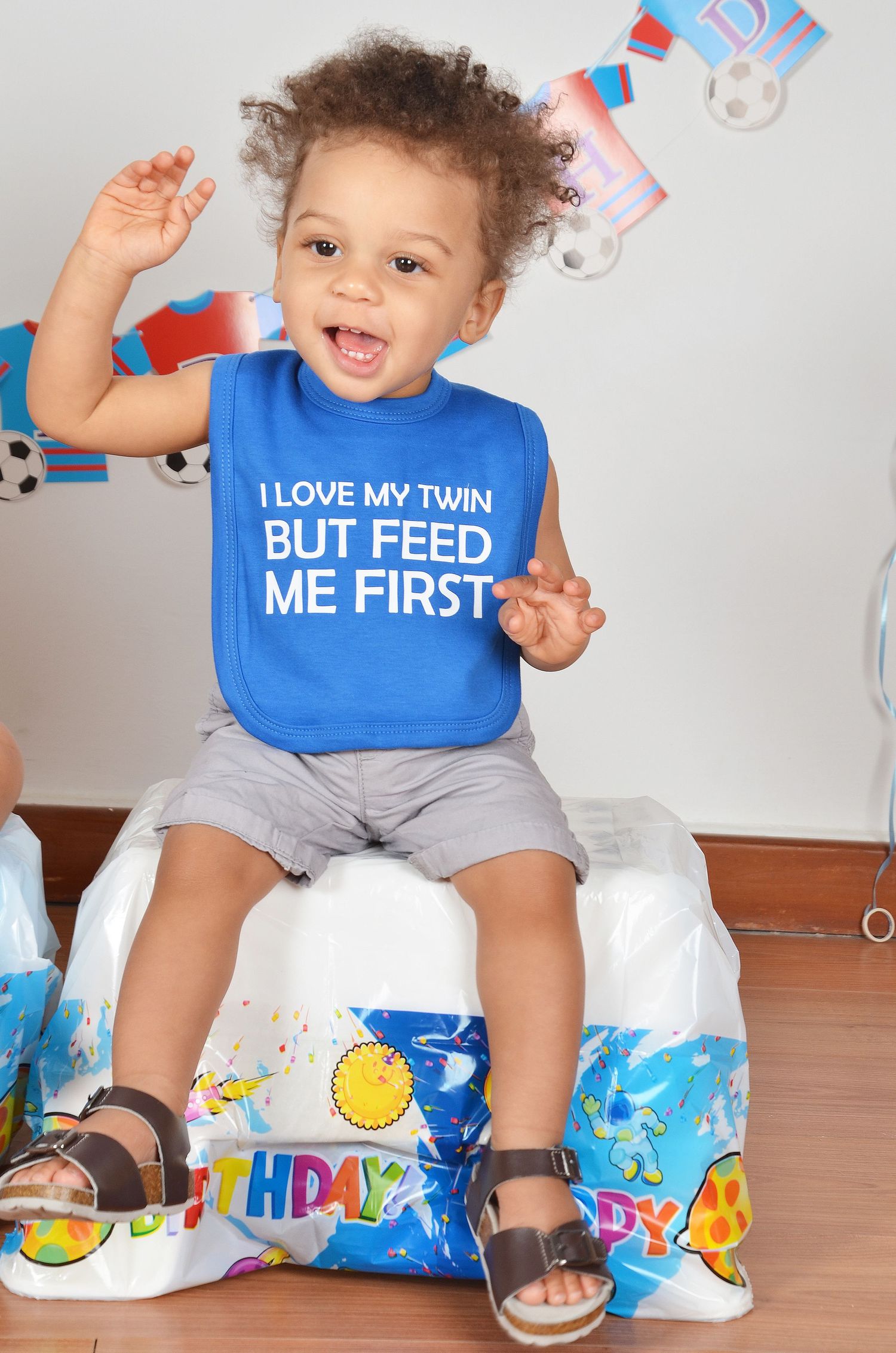A happy toddler in blue bib and shorts sits on birthday decorations with arms raised in celebration.