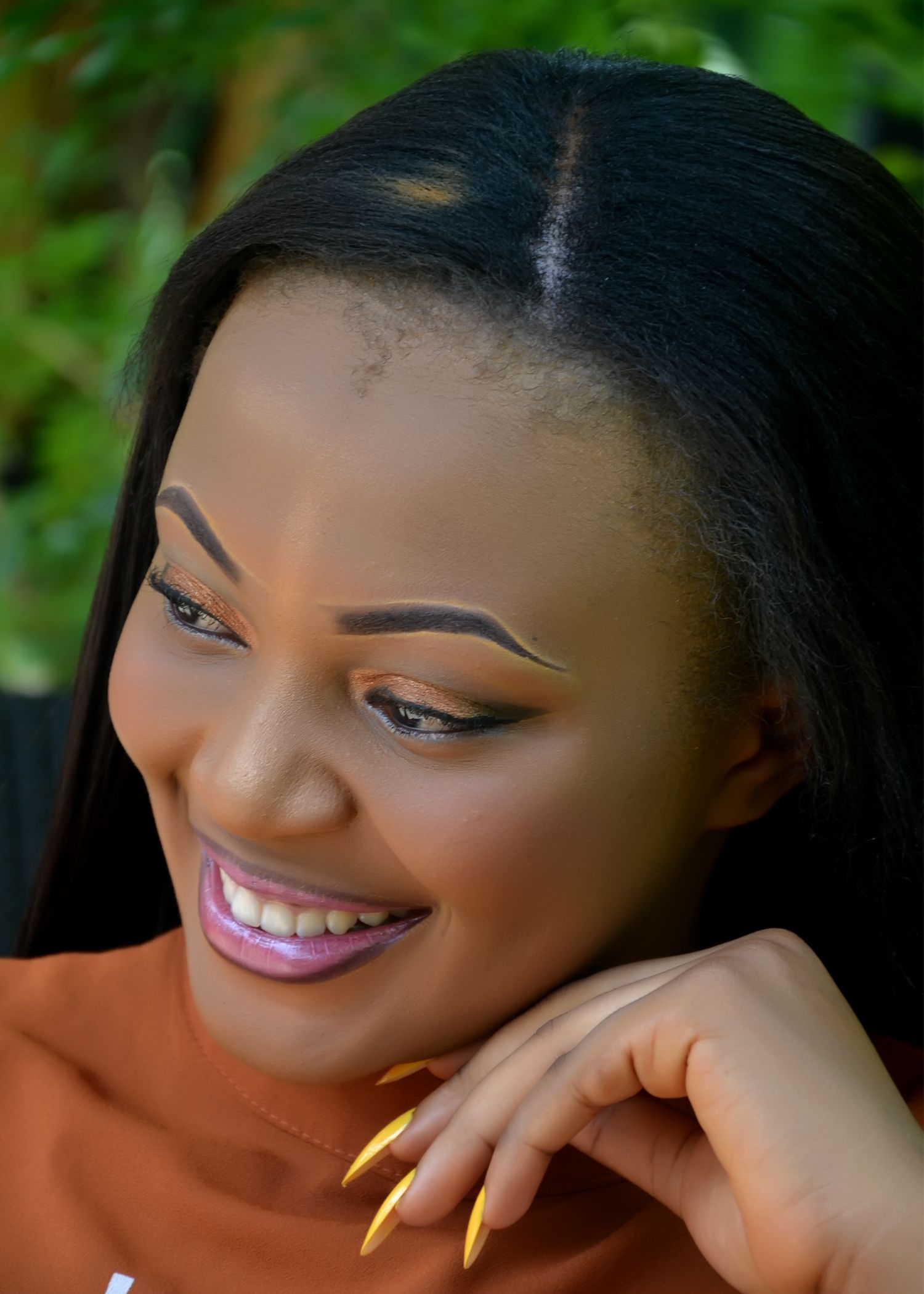 A close-up beauty portrait with yellow nails and mauve lipstick against lush foliage.
