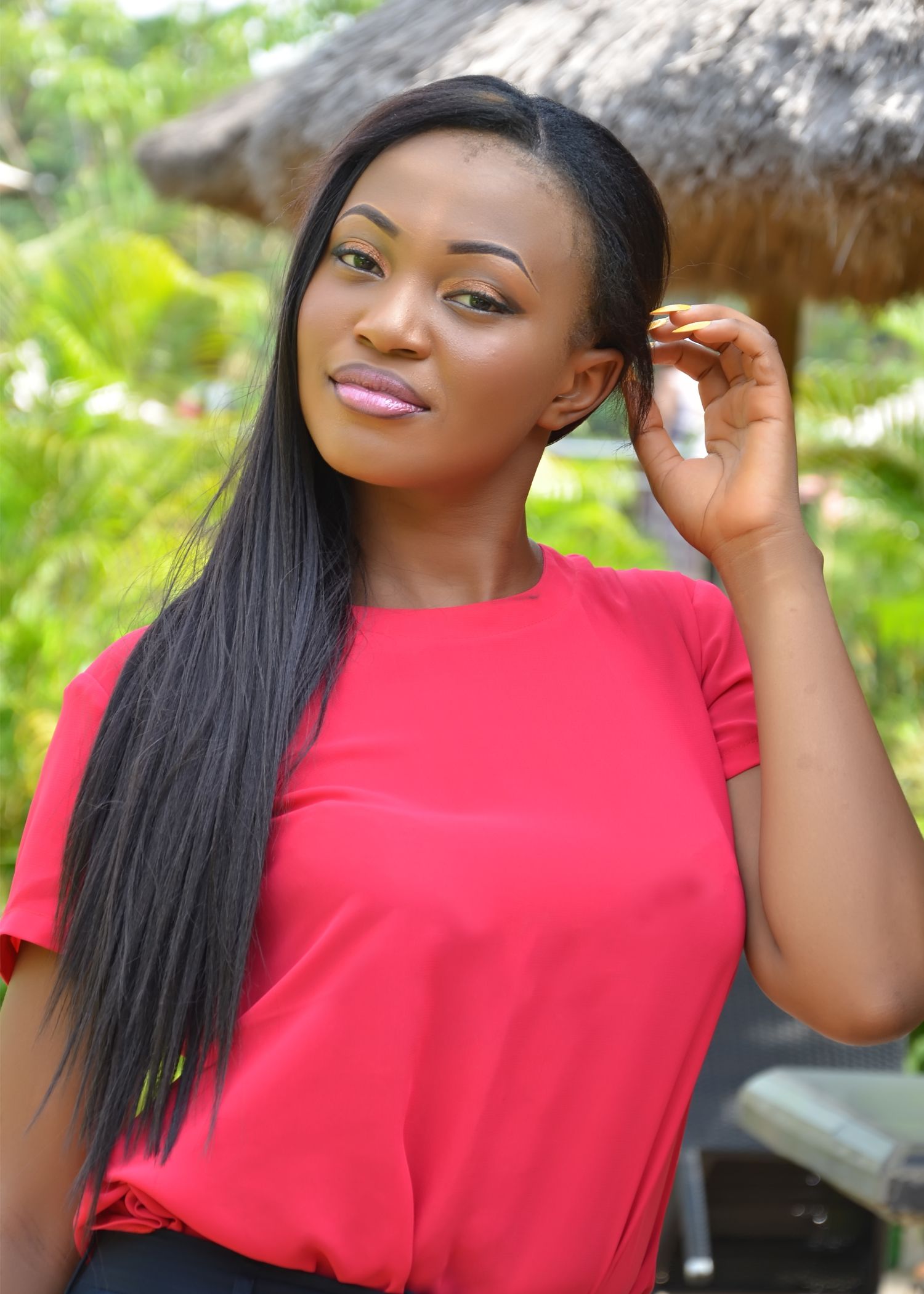 Woman in bright red top poses outdoors against lush tropical background with thatched roof visible.