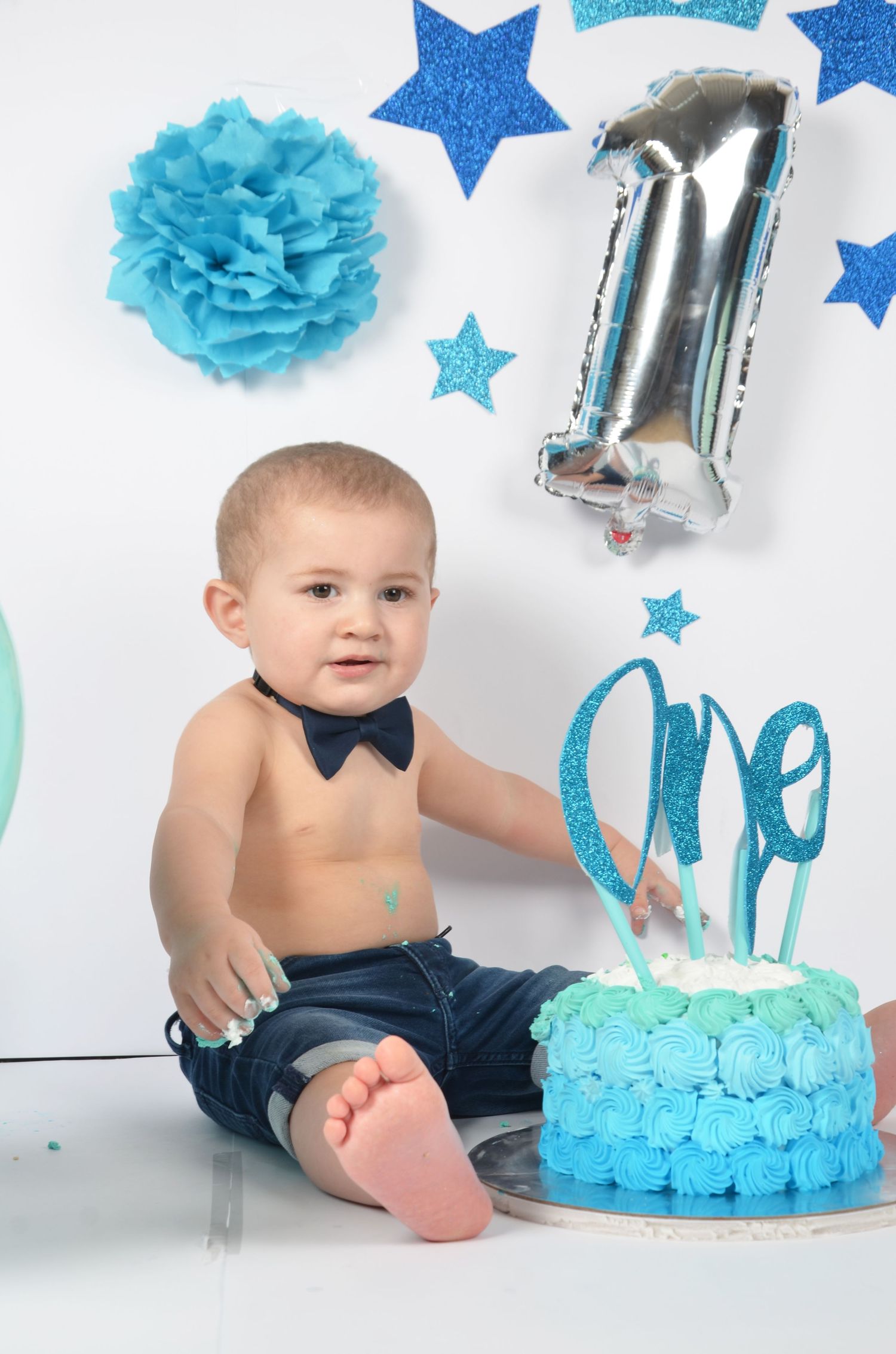 Blue themed first birthday setup with cake, crown decorations, and silver number balloons.