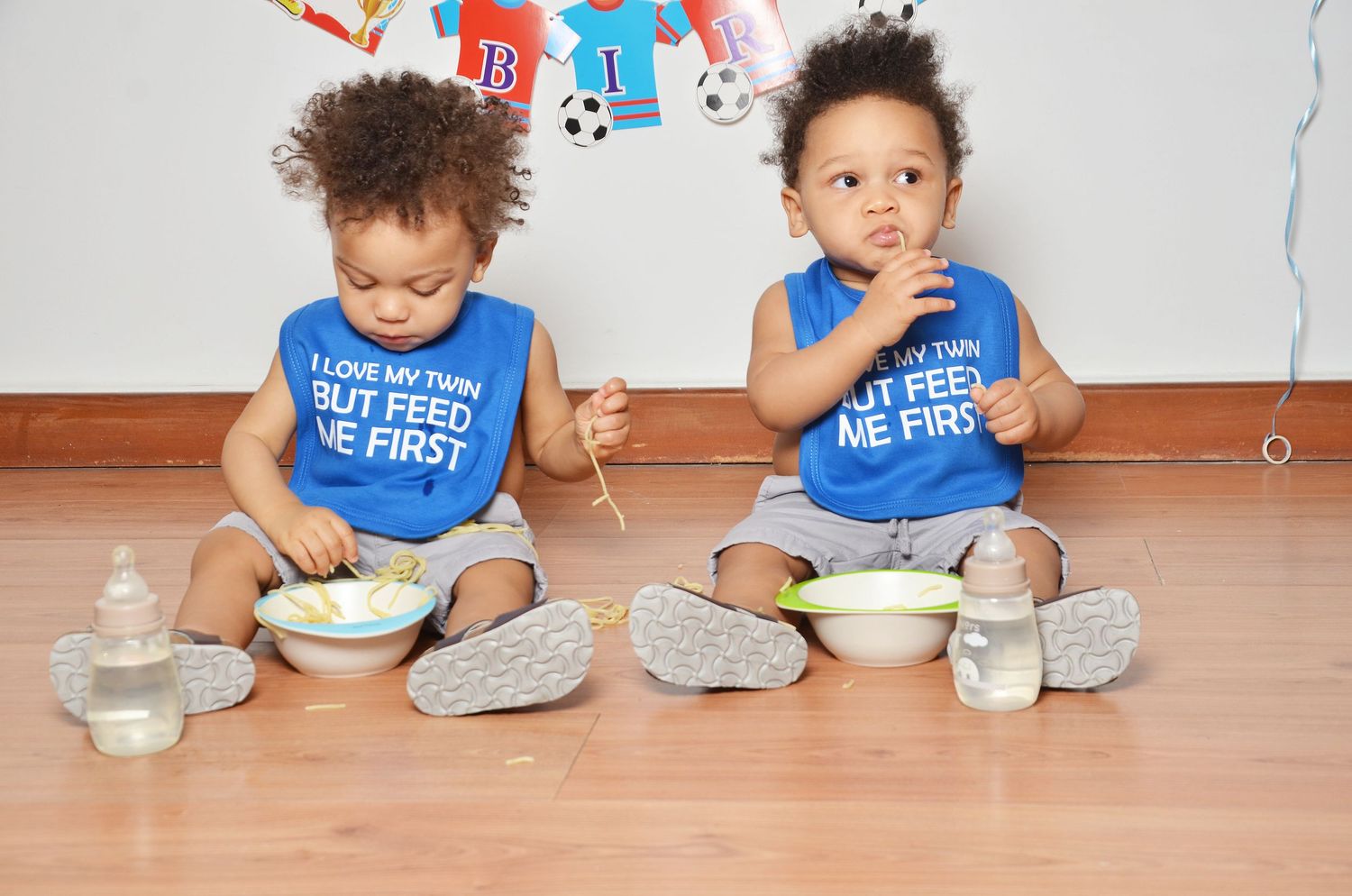 Two babies sitting on hardwood floor wearing matching blue bibs with bottles nearby.