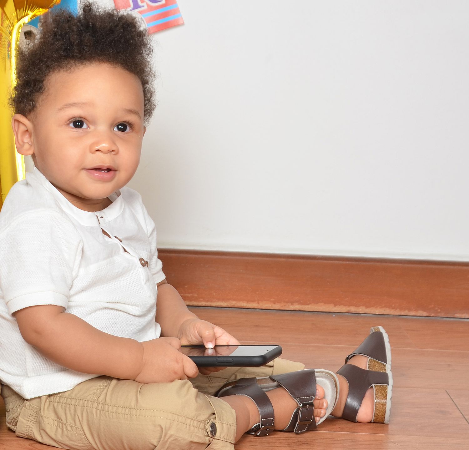Smiling baby in white shirt and sandals sitting on hardwood floor during photo session.