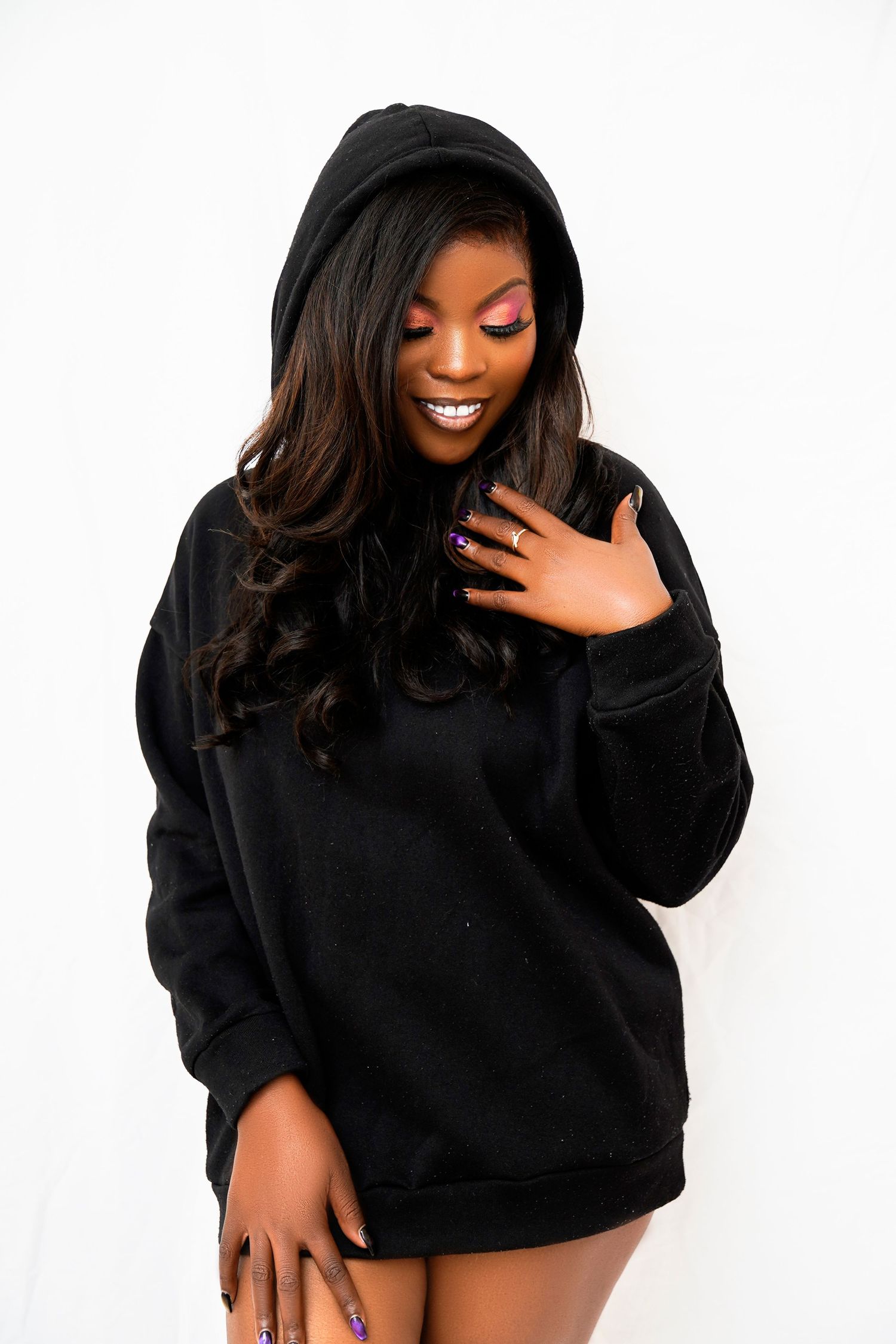 A joyful model in a black hoodie poses against a white backdrop with glamorous makeup.
