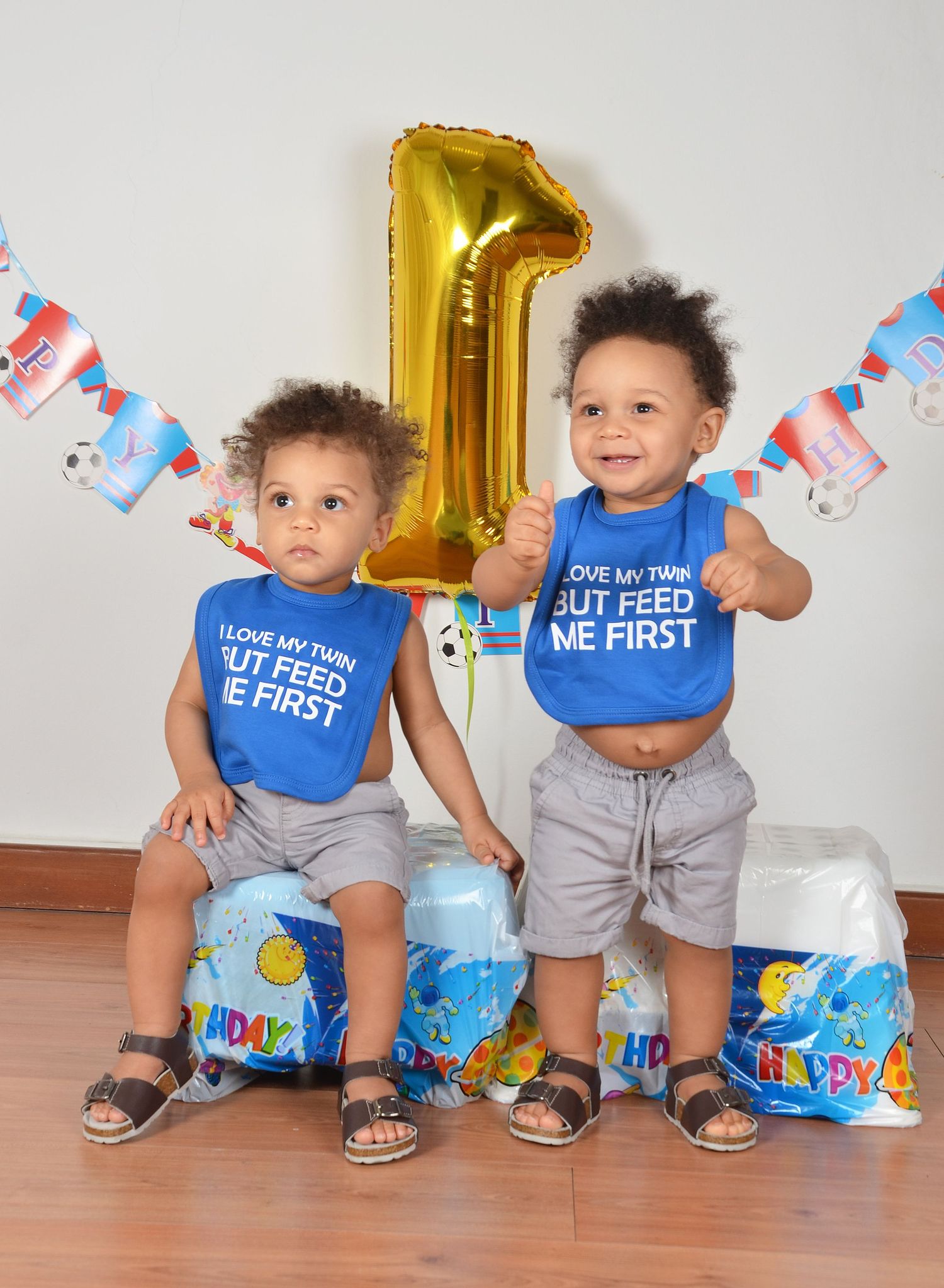 Two toddlers in matching blue shirts sitting together with birthday decorations.