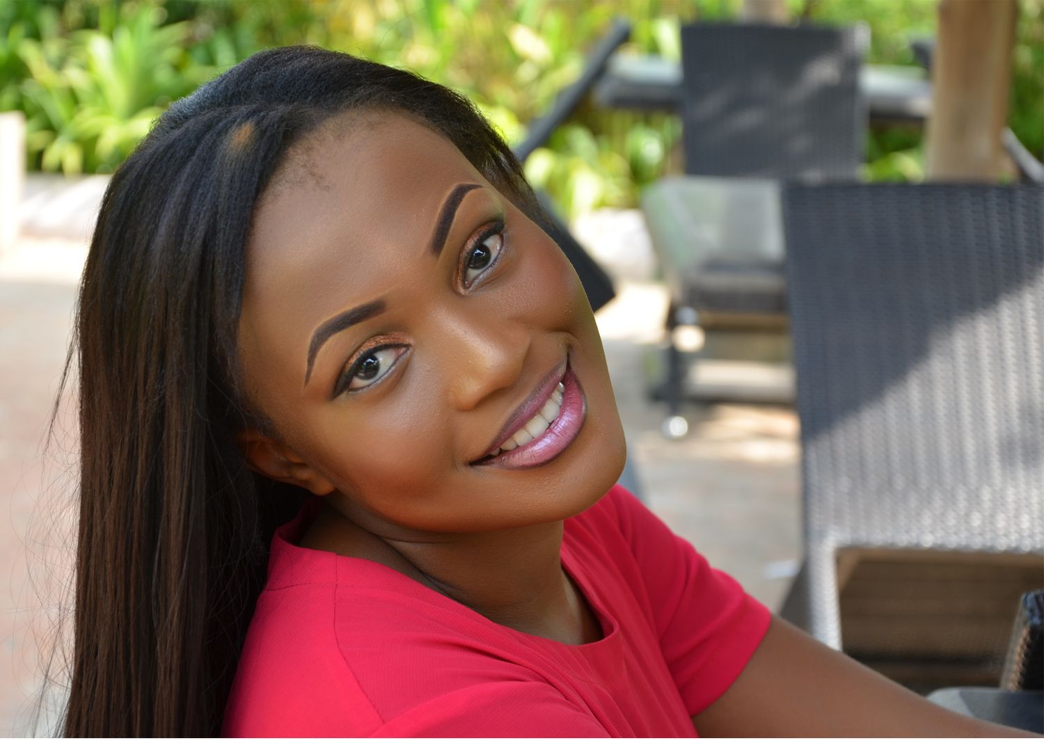 Woman in red top poses casually outdoors with natural lighting and outdoor furniture visible.