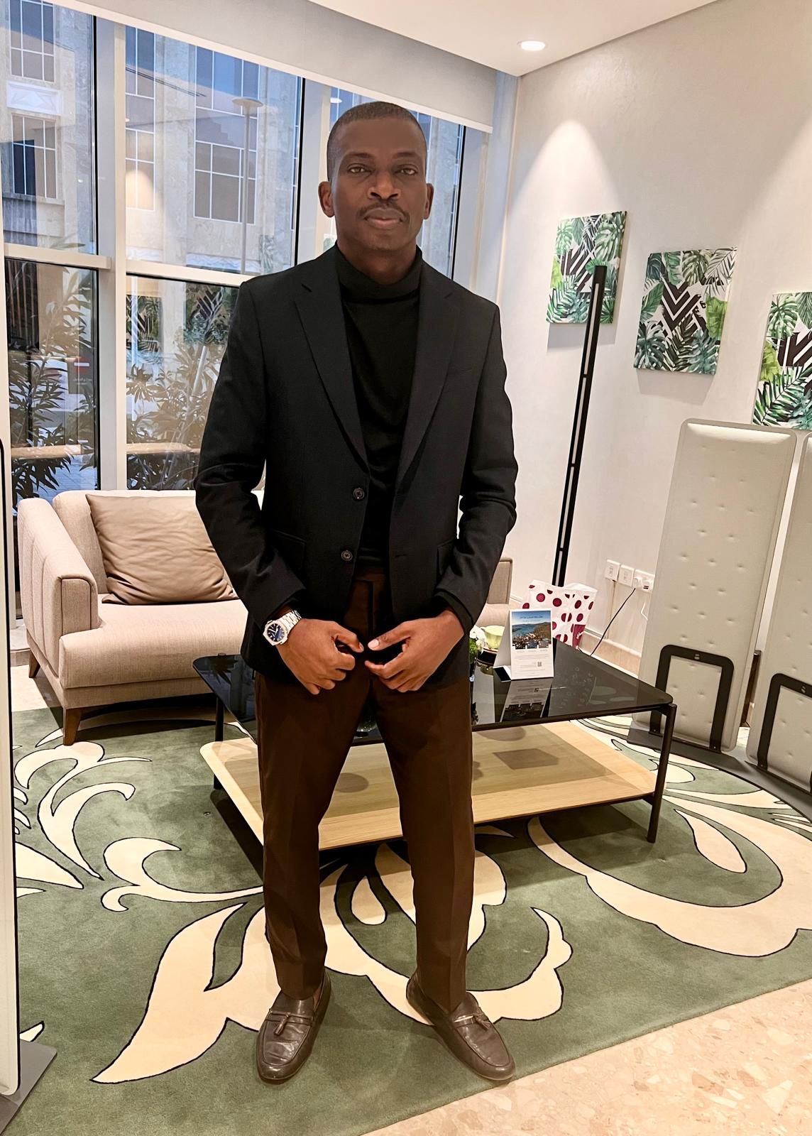 Professional man in black blazer and brown pants standing on decorative rug in modern office setting.