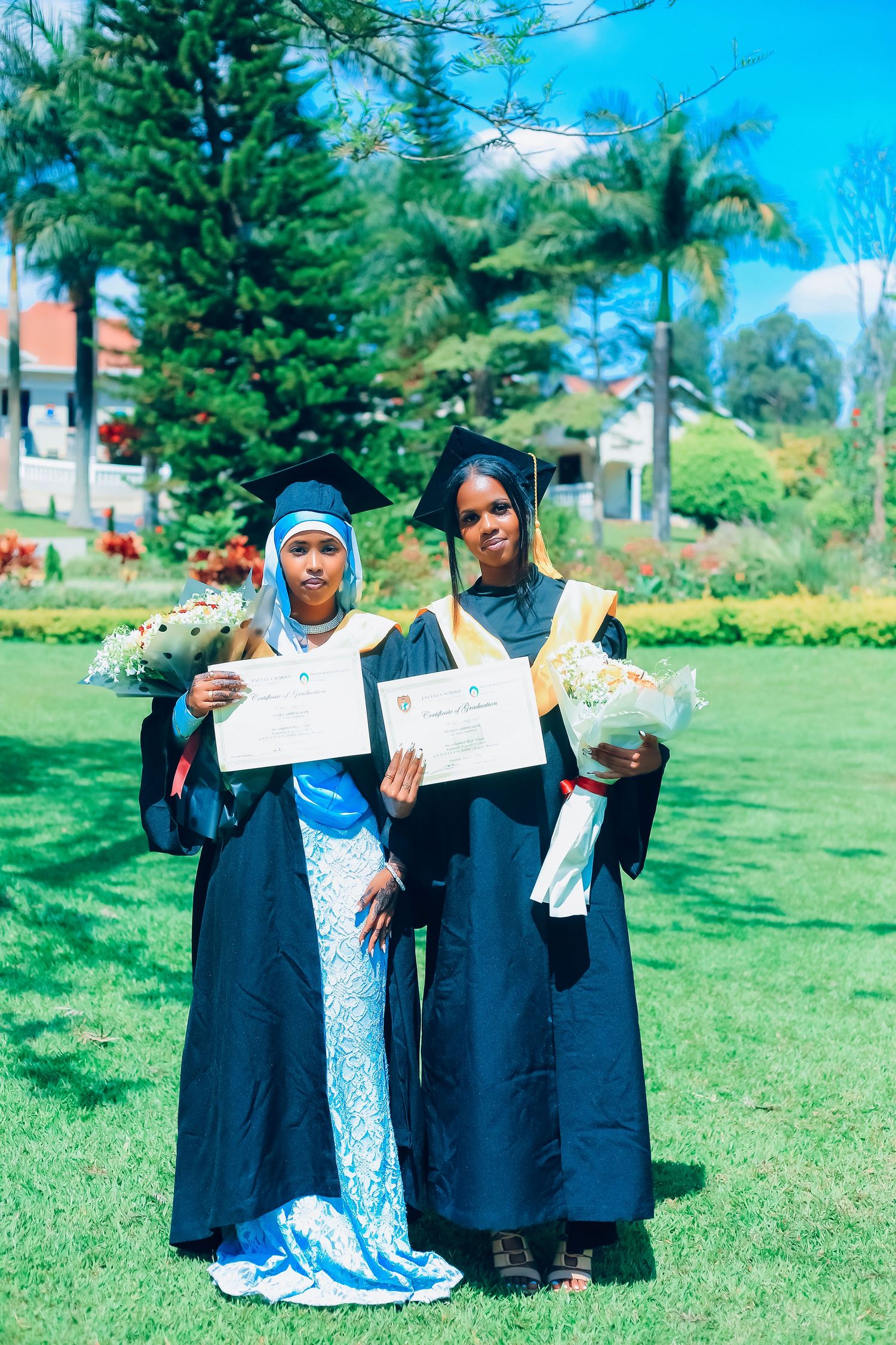 Two graduates in traditional regalia holding diplomas and flowers on sunny campus lawn.