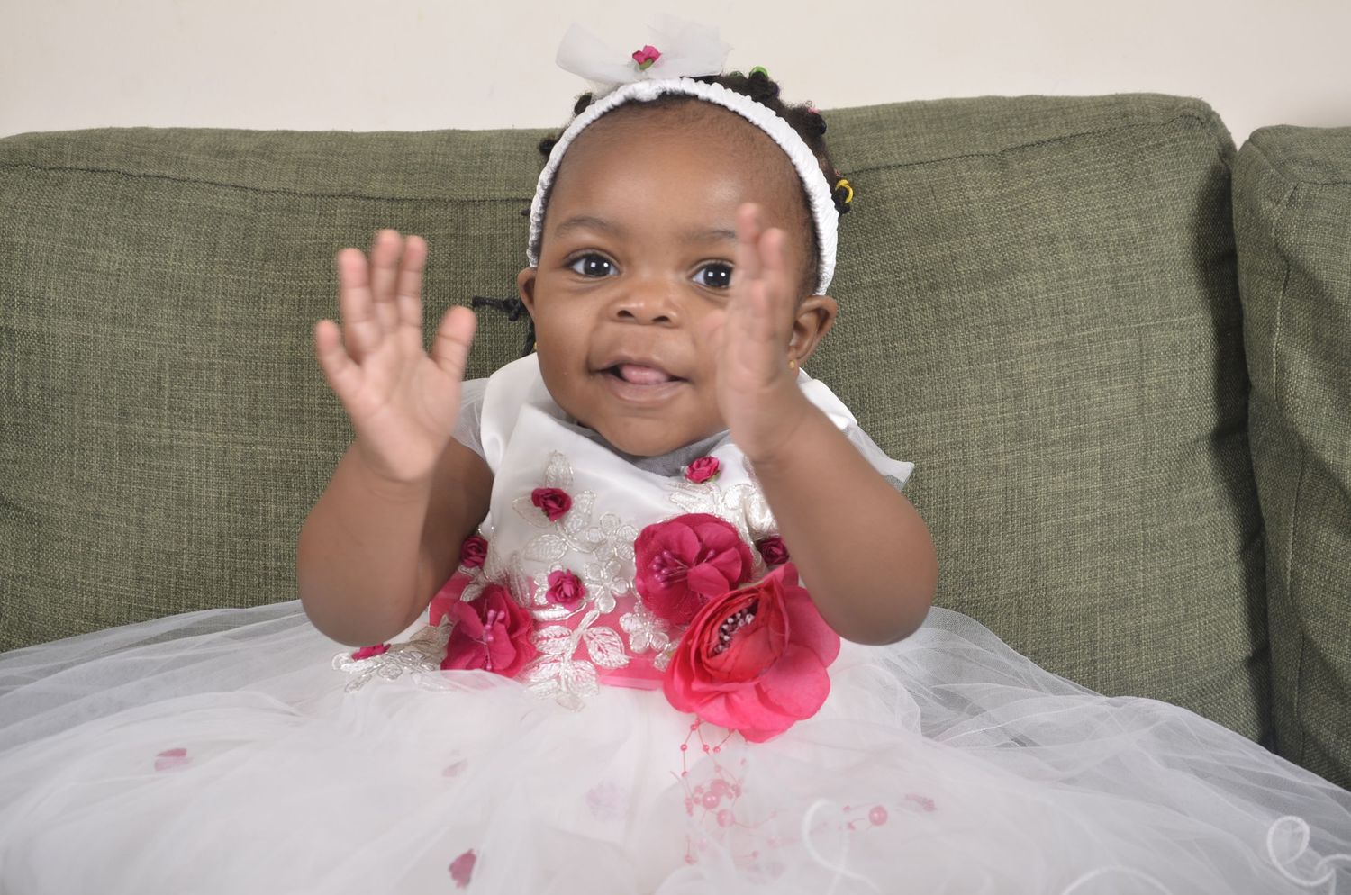 A toddler in a white dress with red floral details seated on a green couch.